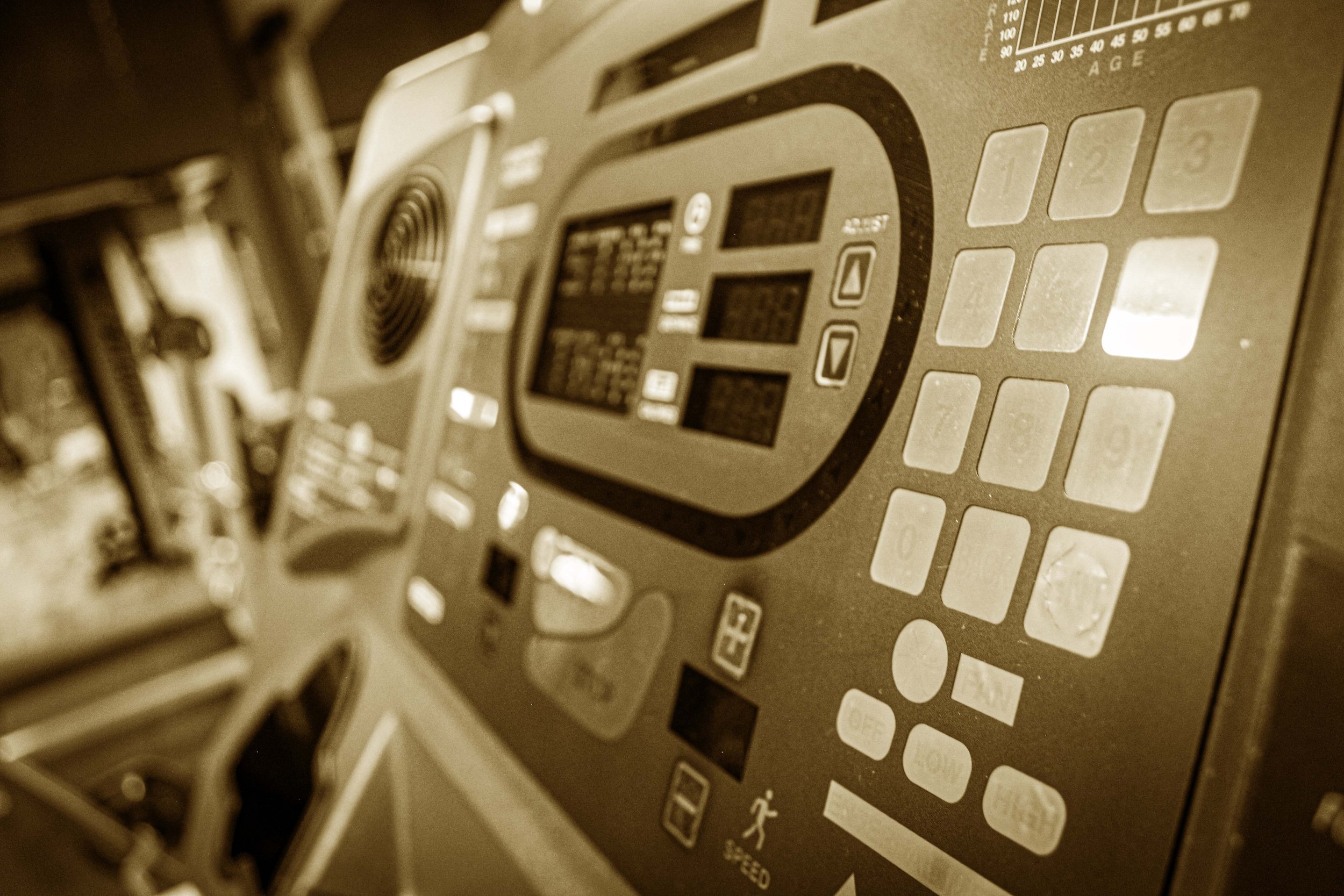 Sepia-toned close-up of a treadmill control panel featuring buttons, a digital display, and various workout settings, with the treadmill frame blurred in the background.
