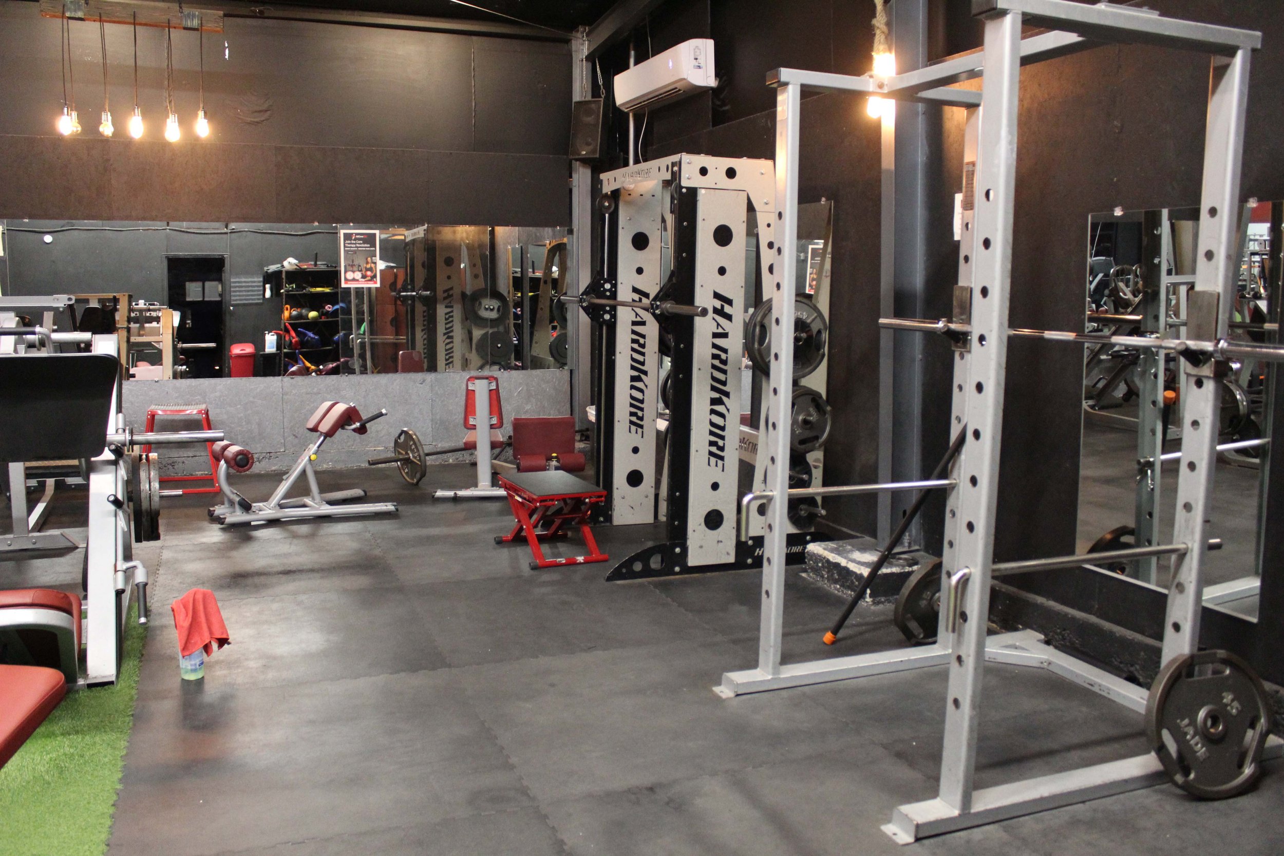 Free weights and squat machines in a gym, featuring a mirrored wall reflecting the equipment, including a HARDKORE squat machine, weight benches, and barbell racks.