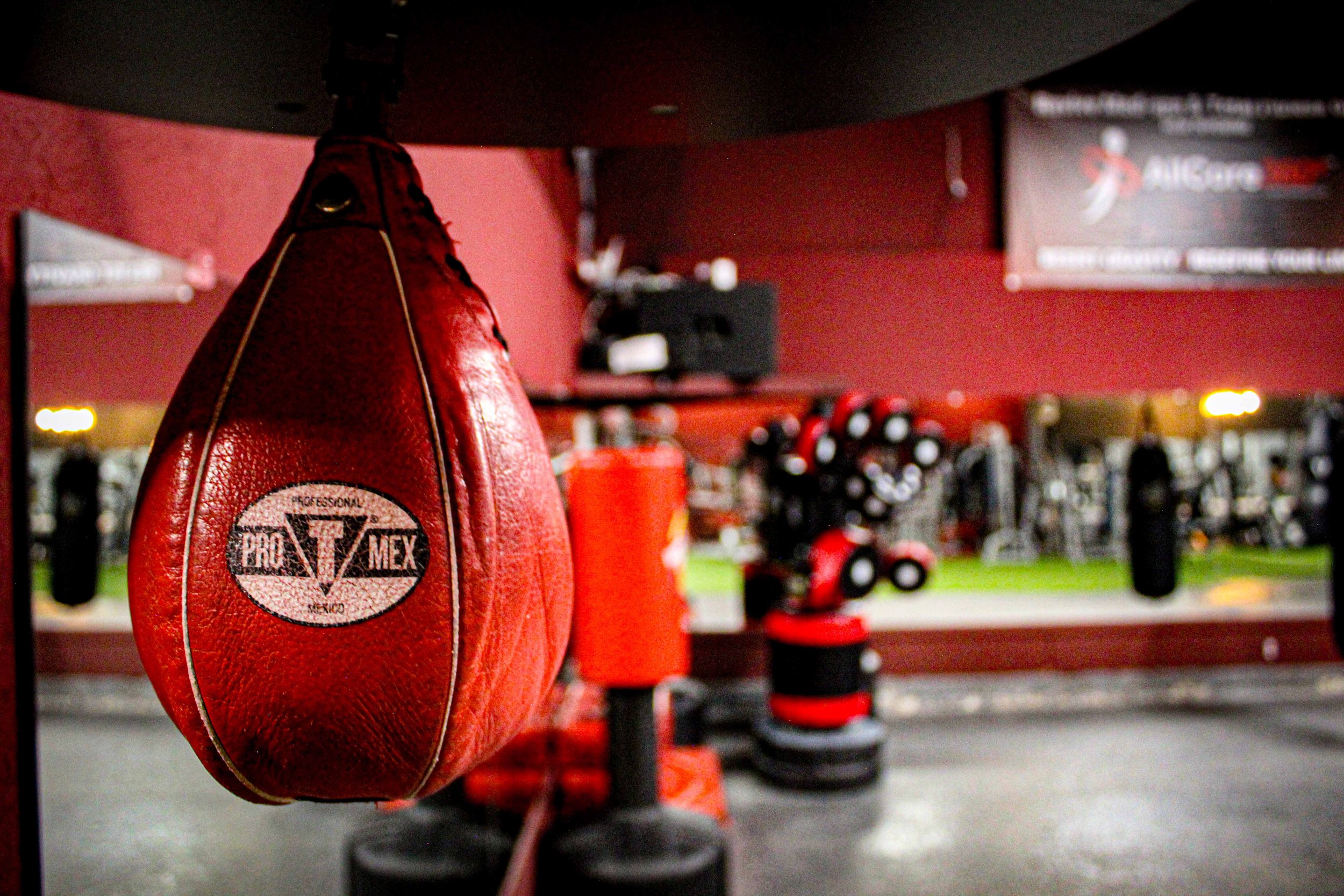 Close-up of a red Pro Mex speed bag in a gym's boxing area, with mirrored walls and red and black training equipment visible in the background