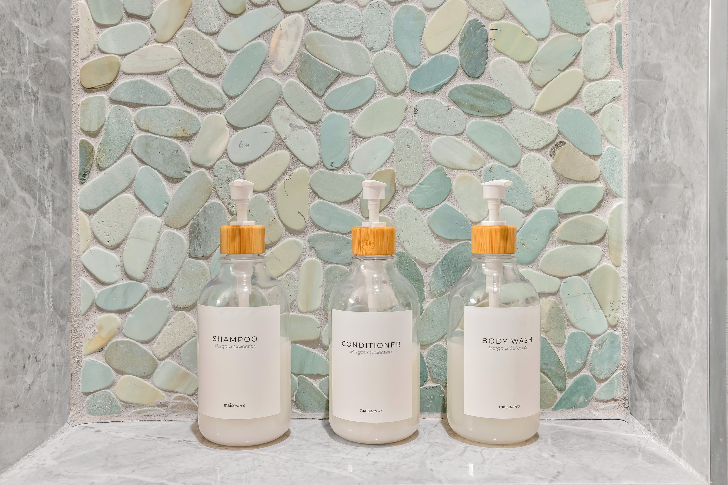 Three clear plastic bottles with wooden caps and white pump dispensers containing shampoo, conditioner, and body wash on a marble shelf in front of a pebble mosaic wall.