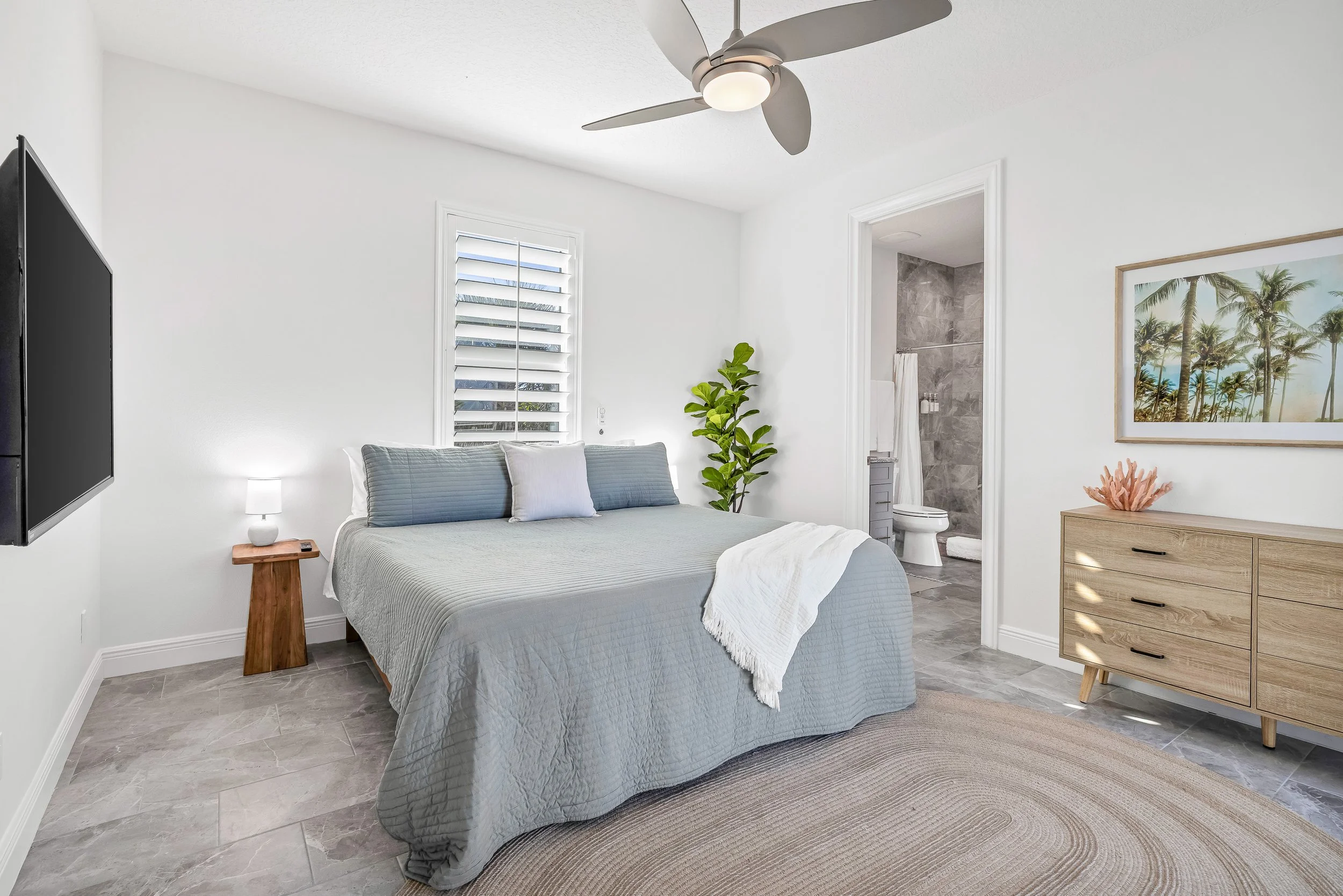 A bedroom with a bed with gray bedding and a white throw blanket, a dark nightstand, a circular beige rug, a white window shutter, and an open door leading to a bathroom with a toilet and shower.