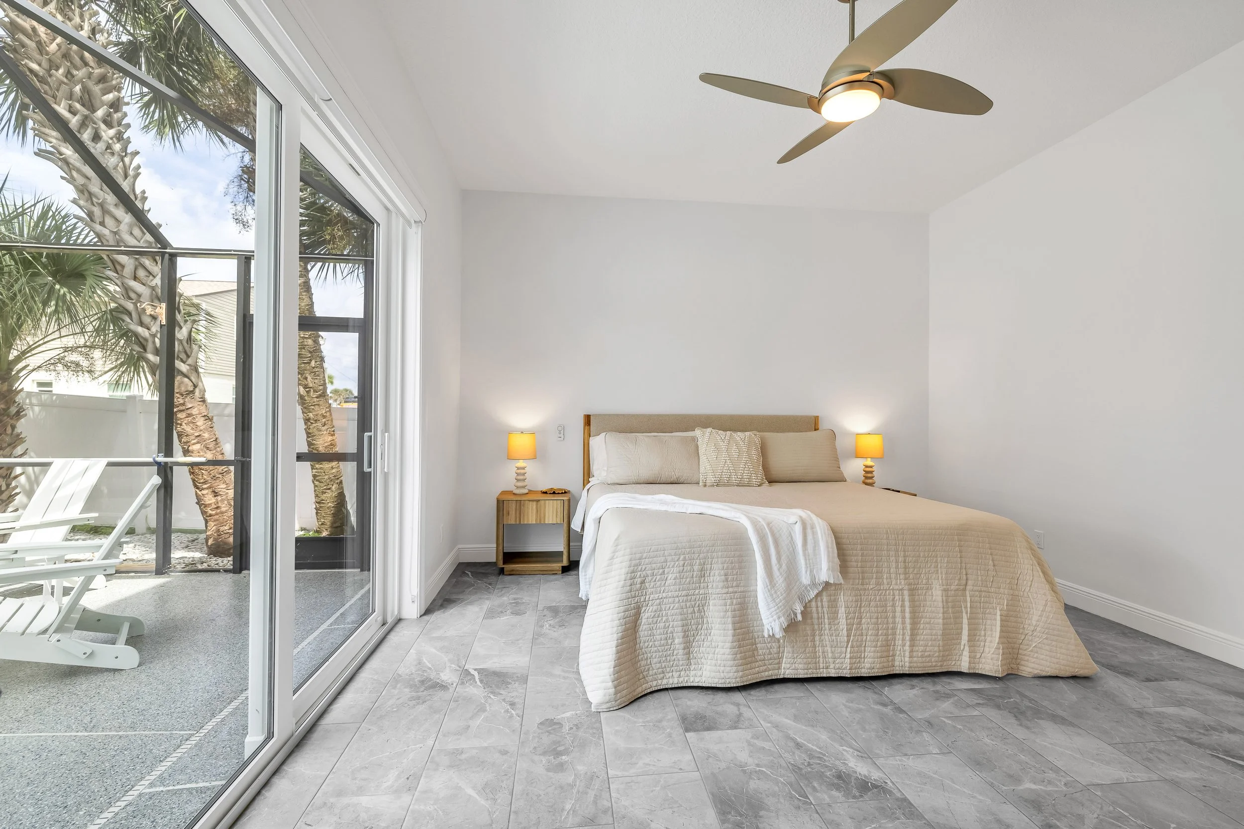 A minimalist bedroom with a large bed, two small bedside tables with yellow lamps, a ceiling fan, and a sliding glass door leading to an outdoor patio with chairs and trees.