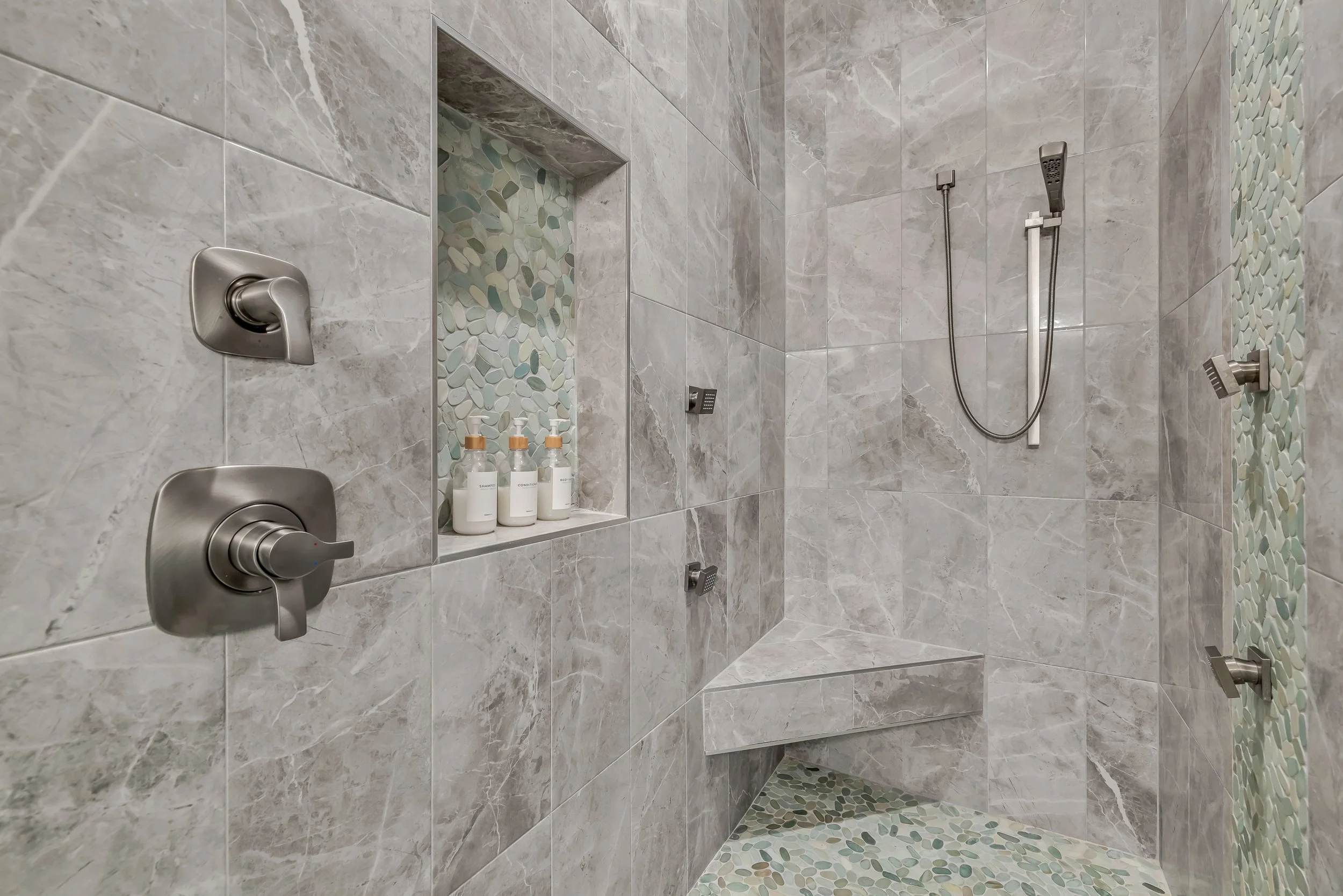 A modern walk-in shower with beige marble tiles on the walls and colored pebble tiles on the floor and a built-in shelf holding bottles of toiletries.