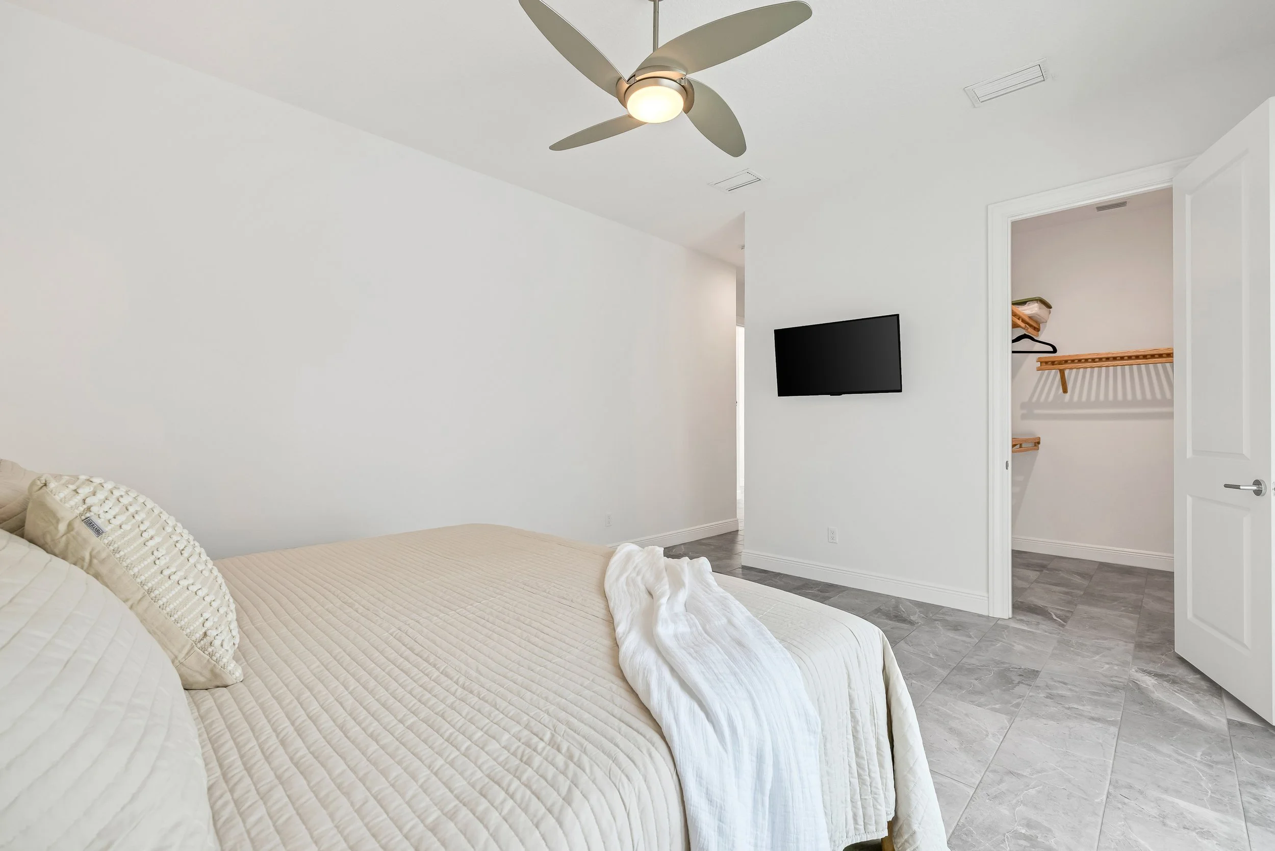 A bedroom with a bed covered in beige bedding and pillows, a ceiling fan, a wall-mounted TV, and a walk-in closet with wood shelving, all in a minimalist style with white walls and light gray tile flooring.
