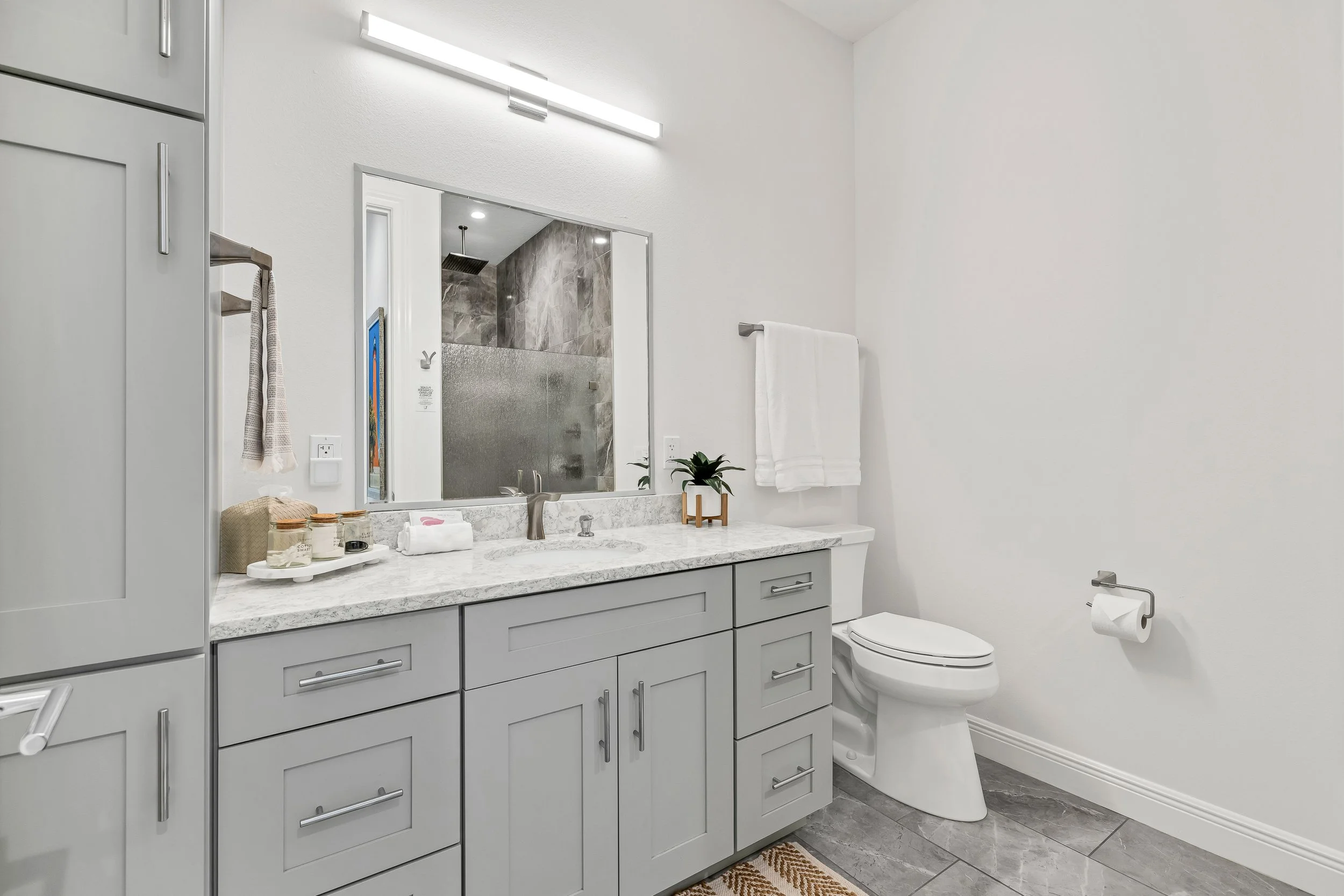 Modern bathroom with light gray cabinets, marble countertop, large mirror, white towel, small plant, and walk-in shower with gray stone tiles.