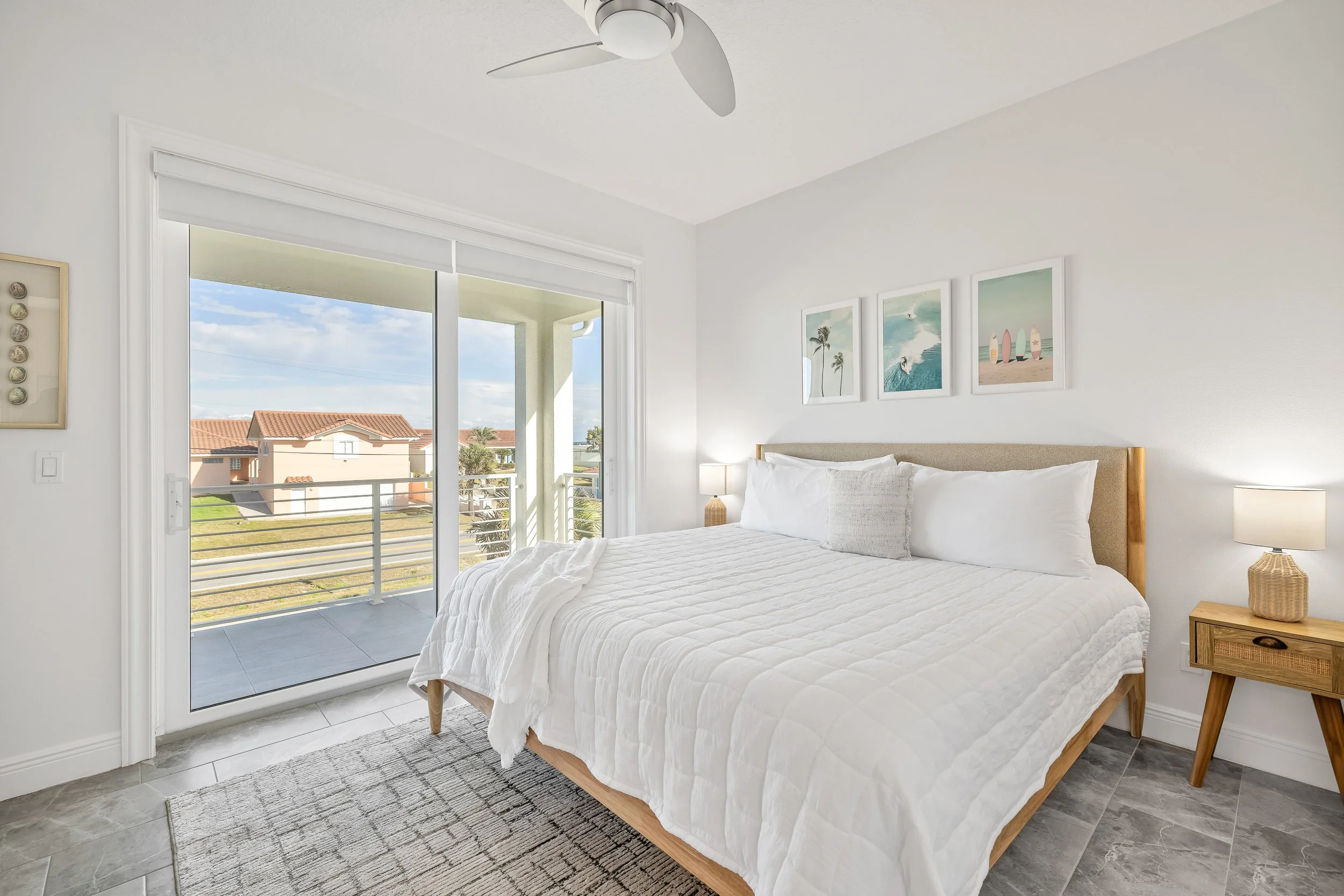 A bright, minimalist bedroom with a large bed covered in white bedding and a beige headboard, a nightstand with a lamp and remote, and a sliding glass door leading to a balcony with outdoor seating and a view of houses and greenery.