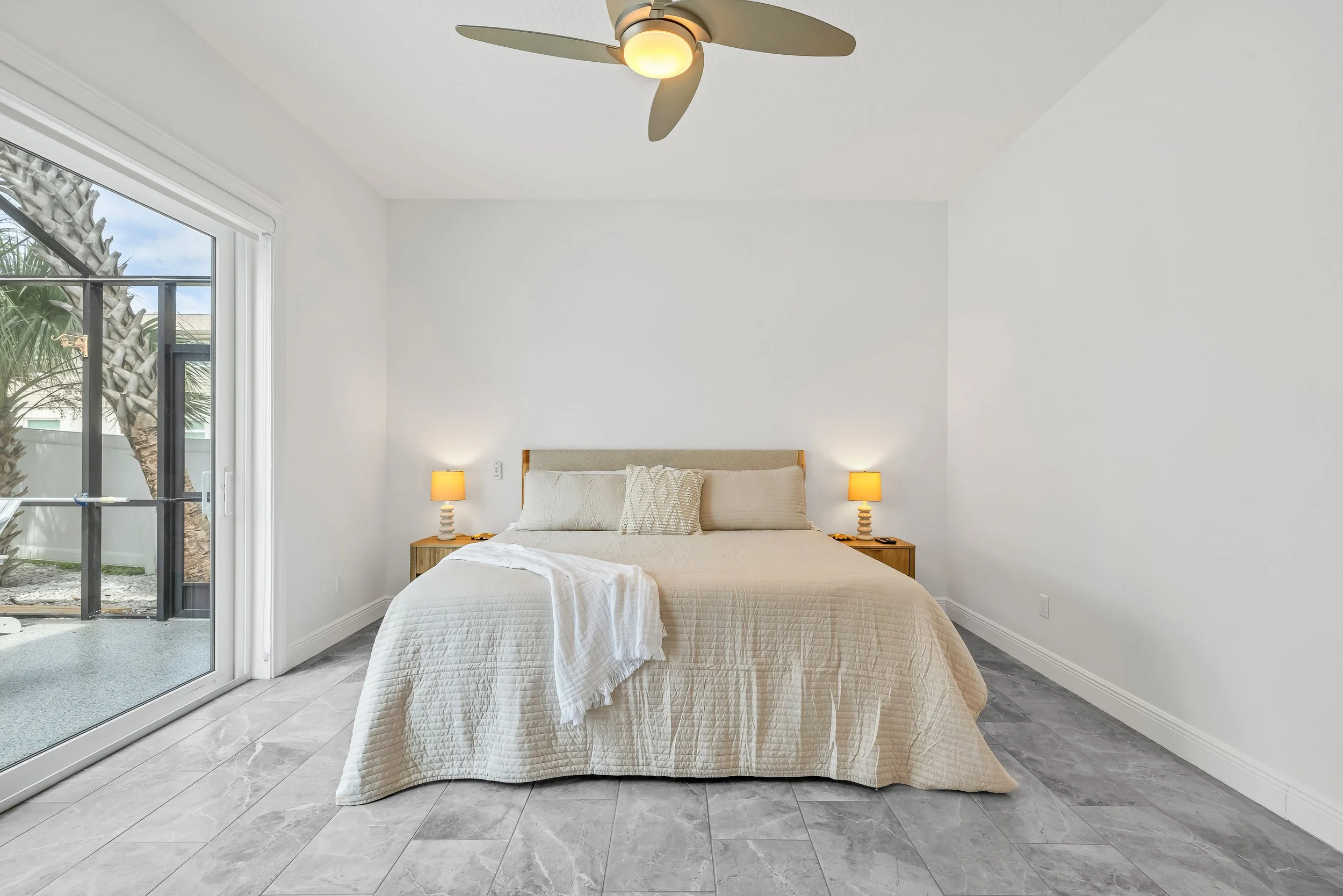 A bedroom with a bed, two nightstands with lamps, and a sliding glass door showing palm trees outside.