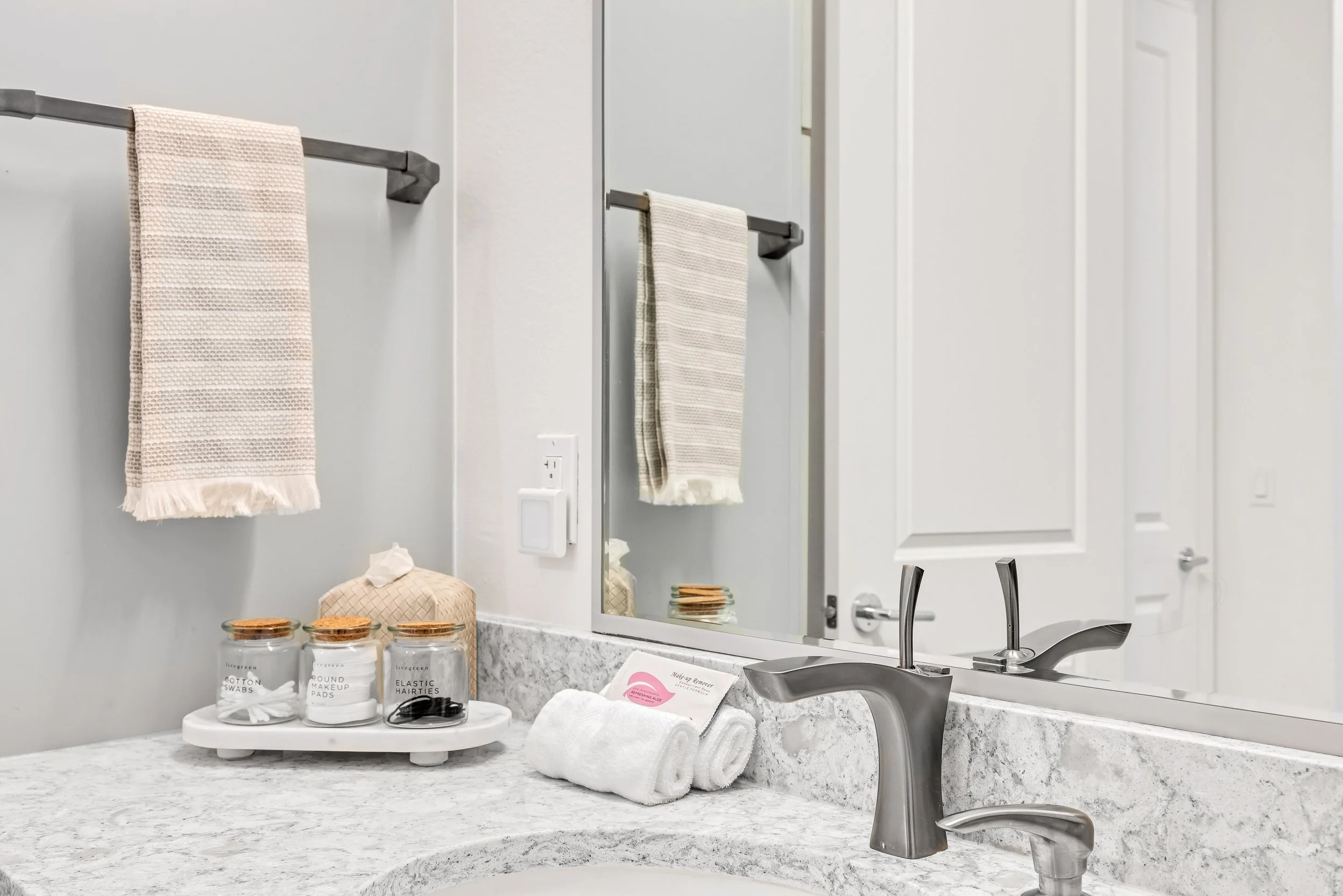 Bathroom vanity with marble countertop, towels, tissue box, jars labeled cotton swabs and makeup pads, and a mirror.