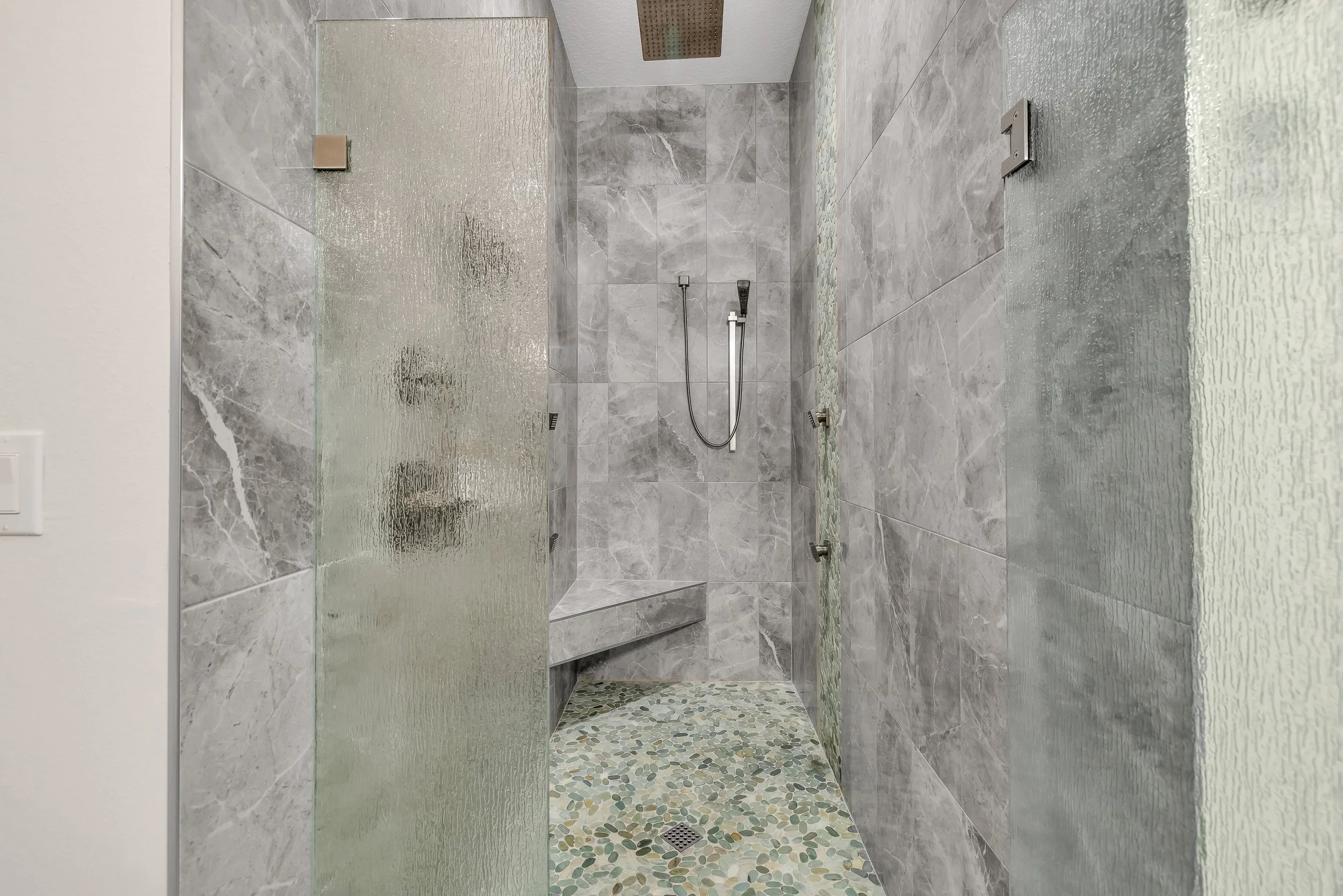 View of a modern walk-in shower with gray marble tiles, a rainfall showerhead, handheld shower, and pebble stone flooring.