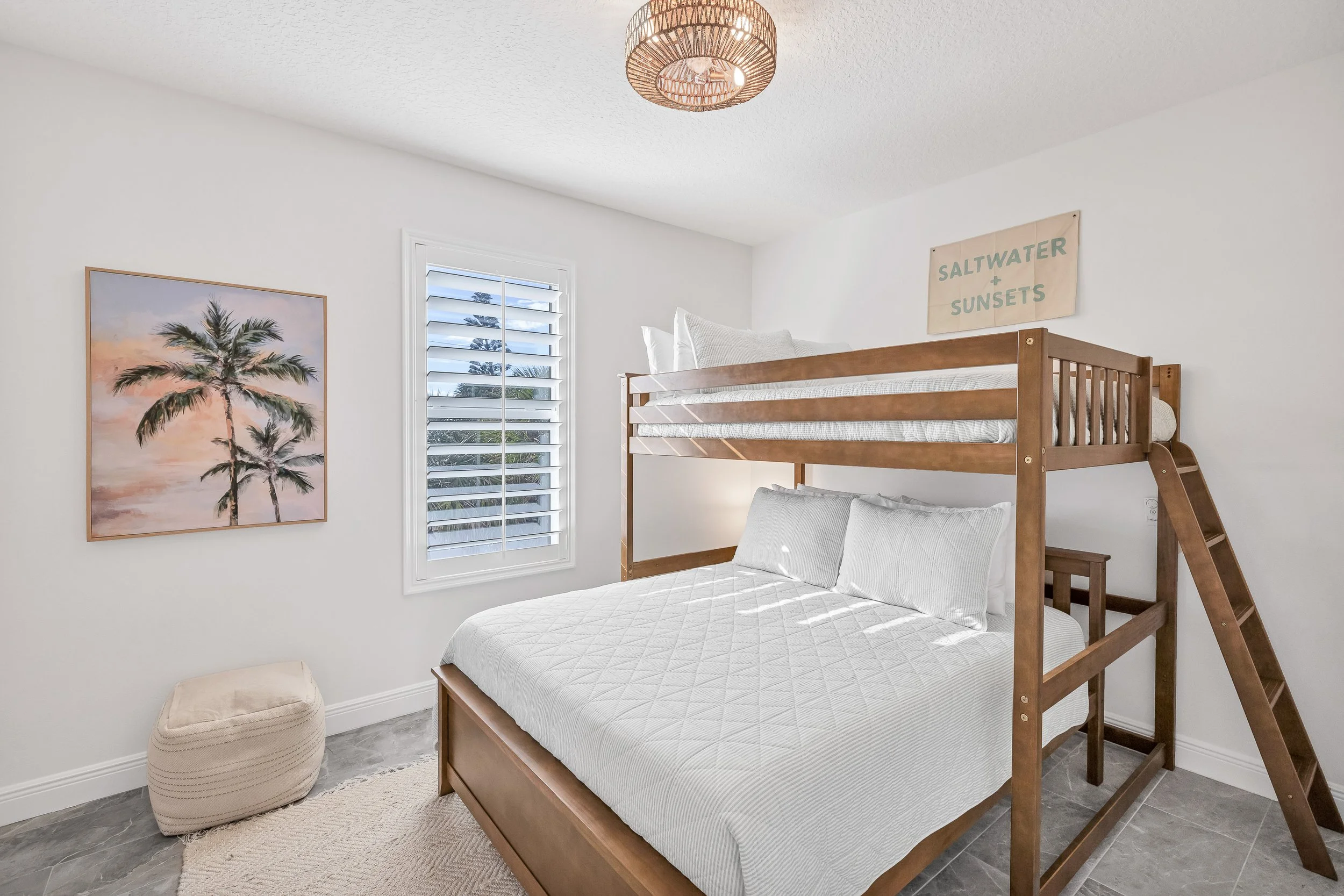 A bedroom with a staircase bunk bed with a lower double bed and an upper twin bed. The room has a window with white blinds, gray tiled floor, white walls, and a hanging light fixture.