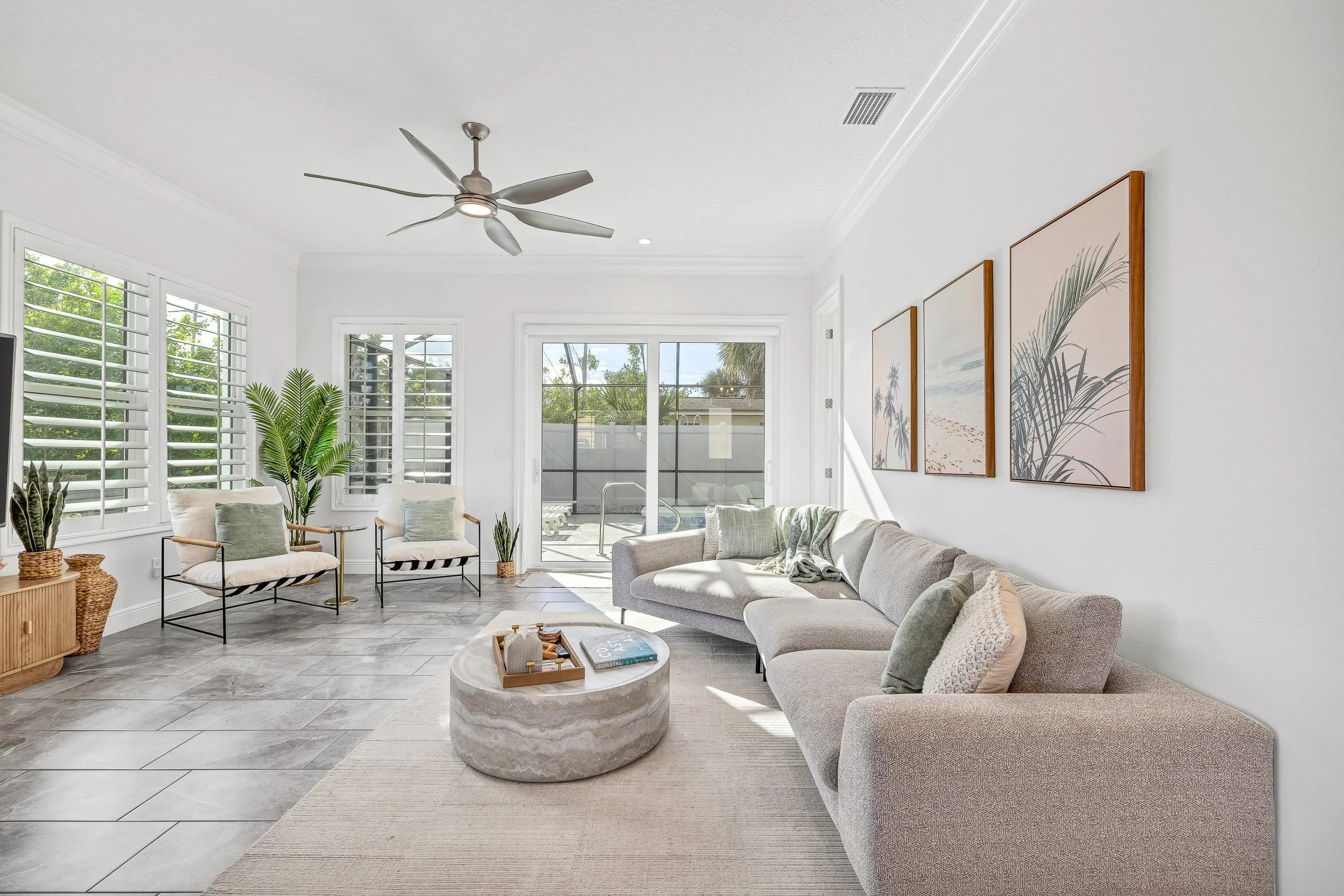 Bright living room with white walls, gray tile flooring, a gray sofa with pastel pillows, round beige coffee table, white armchairs with pastel cushions, large windows with plantation shutters, and sliding doors leading to an outdoor pool area.