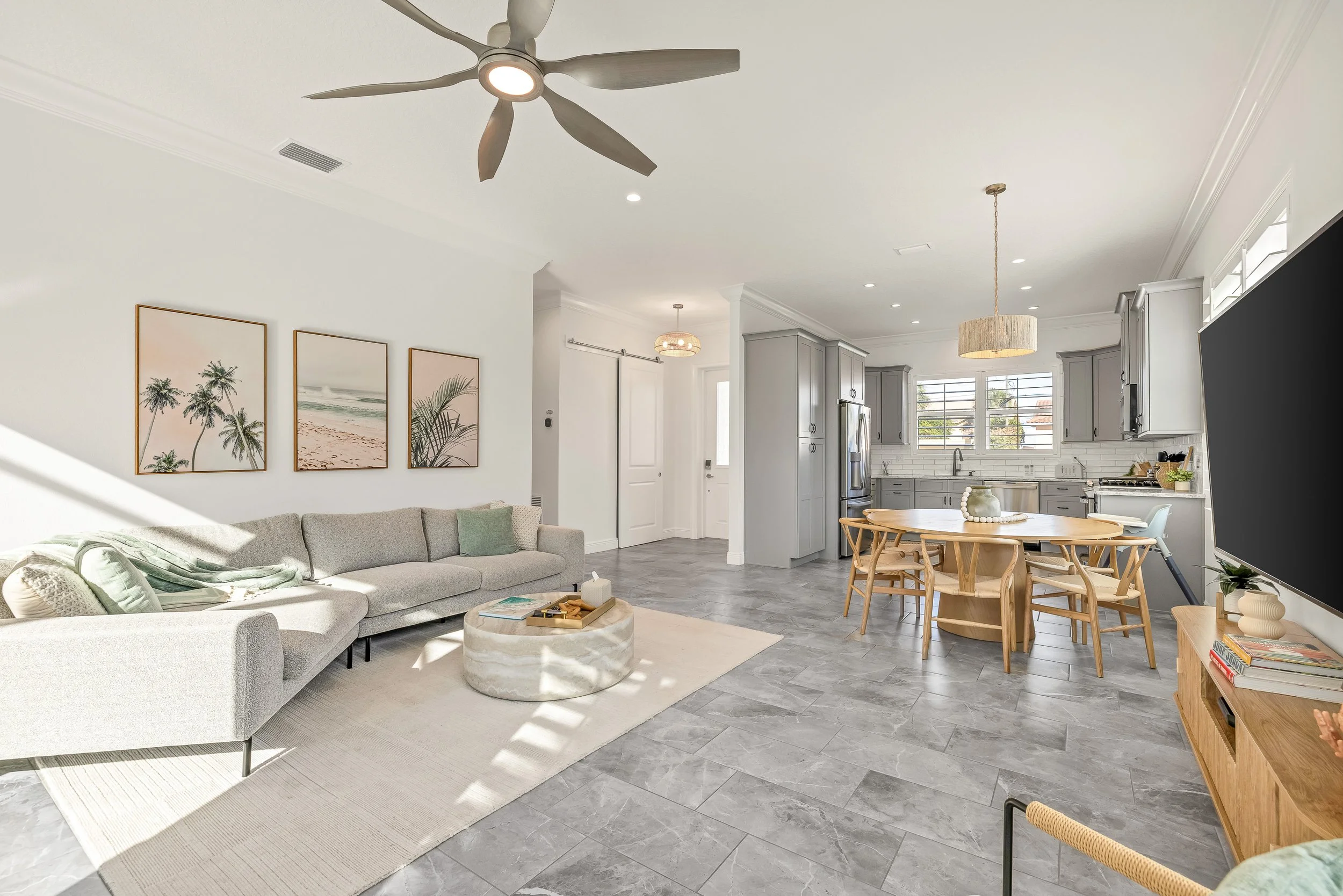 Bright and airy open-concept living room and kitchen with gray and white color scheme, large windows with white shutters, modern ceiling fan, and minimalist decor.