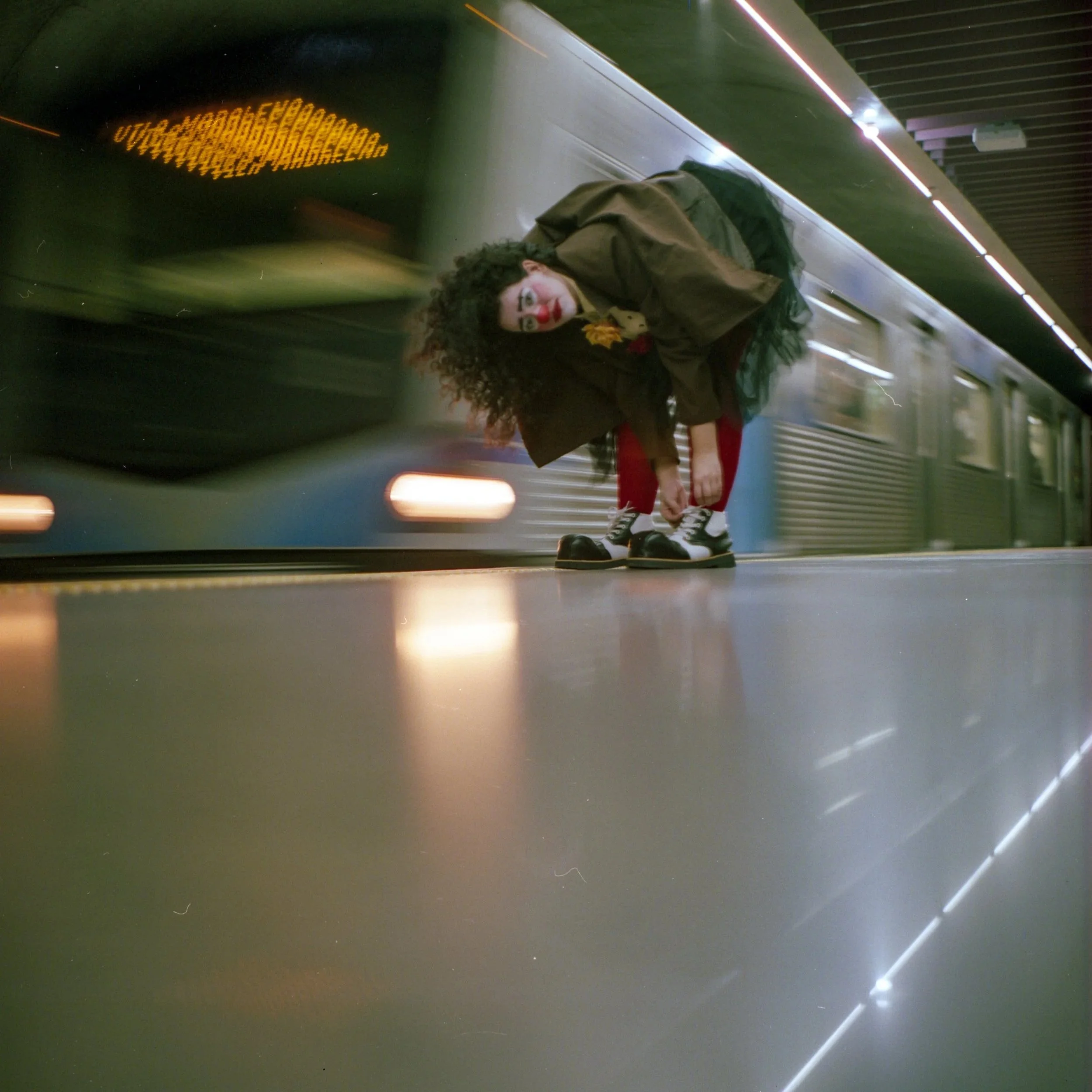 A woman with curly hair crouching on a subway platform, tying her shoelaces, with a moving train behind her.