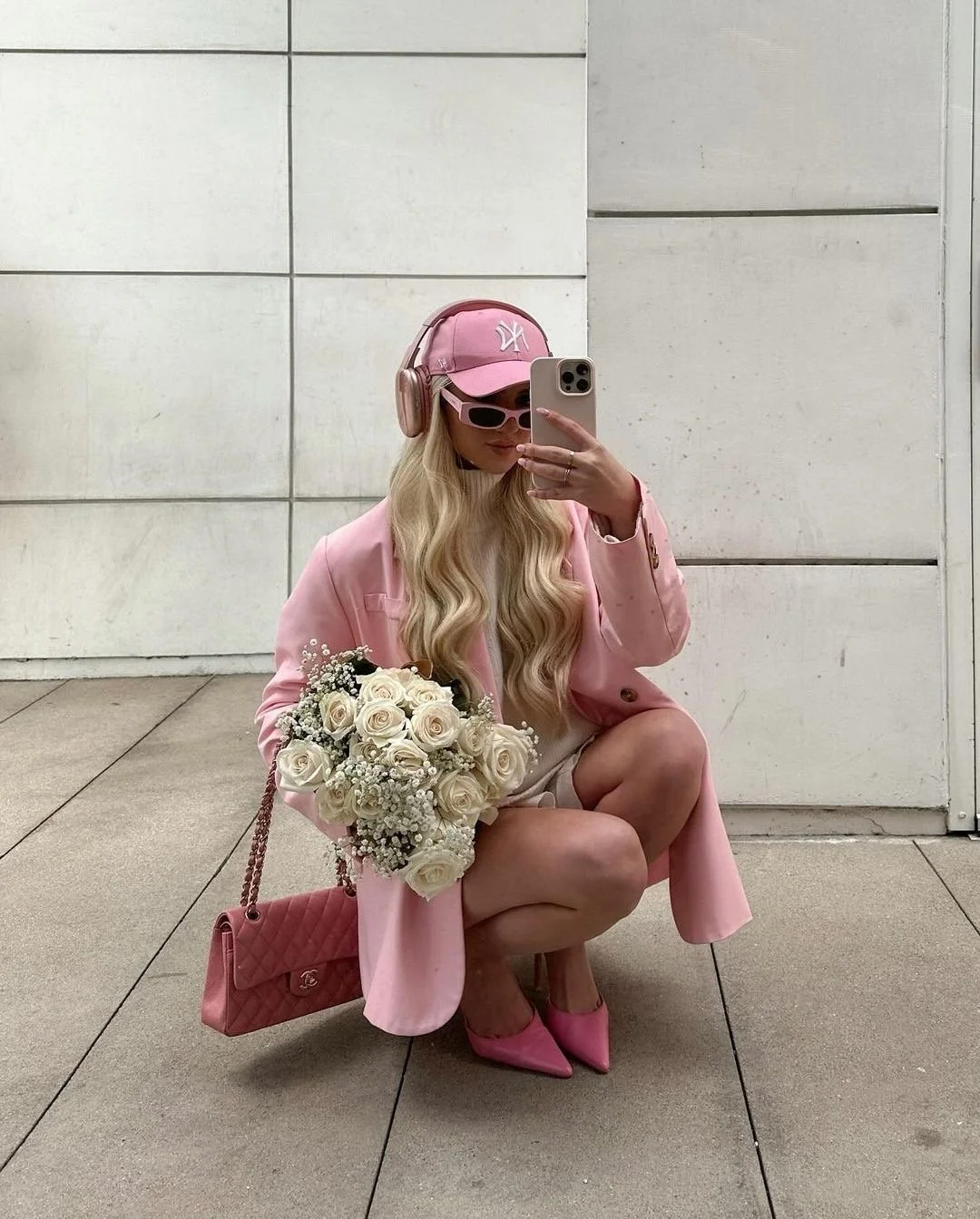 Woman squatting for a mirror selfie wearing a pink coat, pink baseball cap, and sunglasses, holding a bouquet of white roses and a pink quilted handbag.