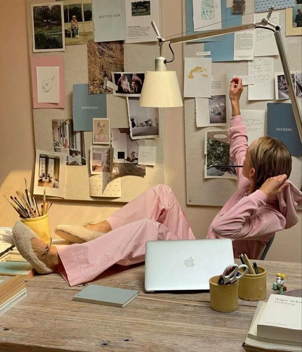 Artist wearing a pink lounge set relaxing in a creative studio with feet on the desk, looking at a large inspiration mood board filled with art prints.