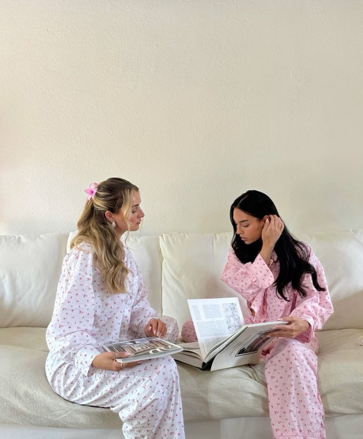 Two friends sitting on a white sofa wearing matching floral print pajamas, reading fashion magazines and talking.