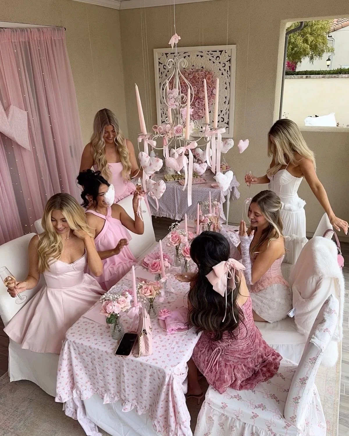 Group of friends in pink and white dresses celebrating at a pink-themed party with floral table decorations, candles, and hanging heart ornaments.