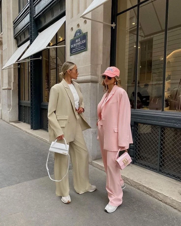 Two women posing on a city street wearing oversized suits in beige and pink, styled with sneakers, sunglasses, and mini handbags.