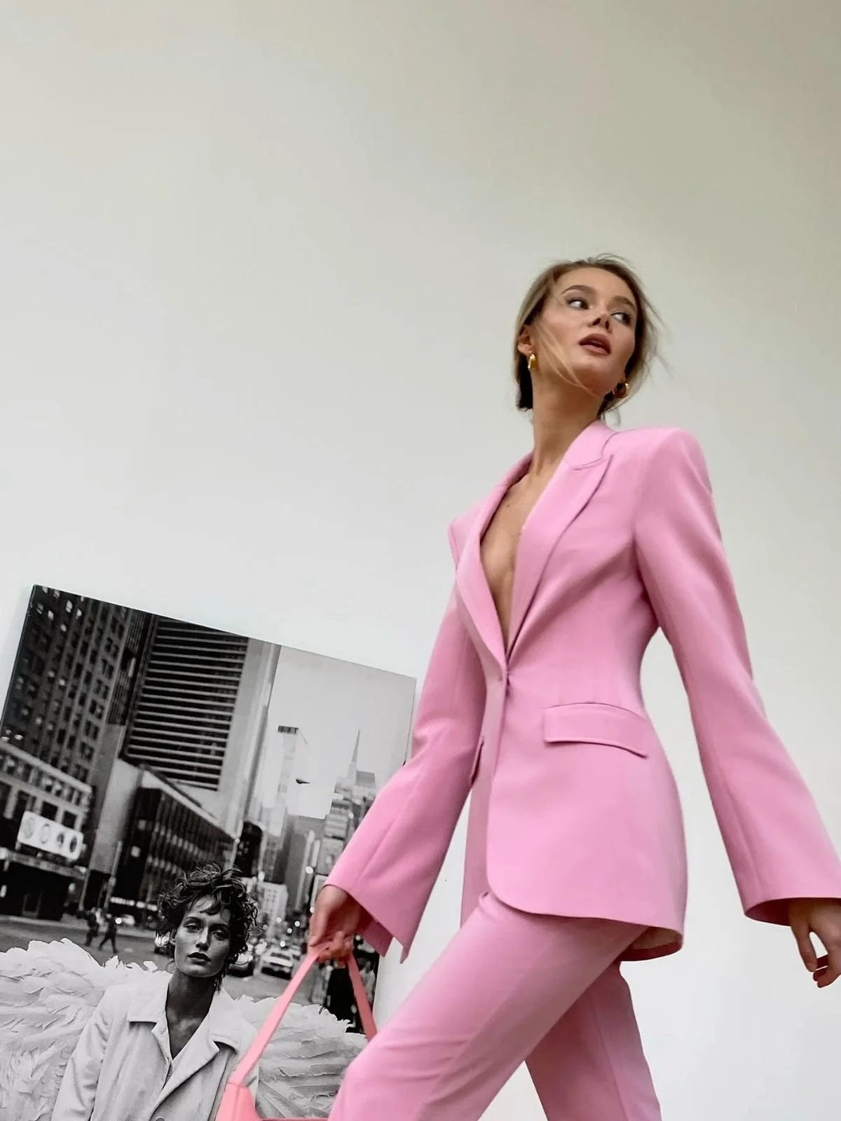 Low-angle fashion shot of a woman walking in a tailored pink suit and matching trousers, carrying a pink bag, with a black and white city art print in the background.