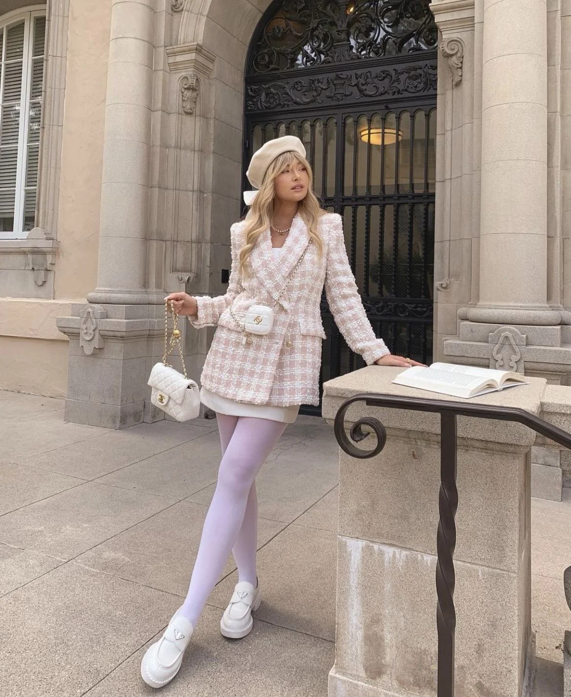 Fashion photo of a woman posing outside a stone building wearing a pink tweed blazer, white beret, white tights, and loafers, holding a quilted white handbag.