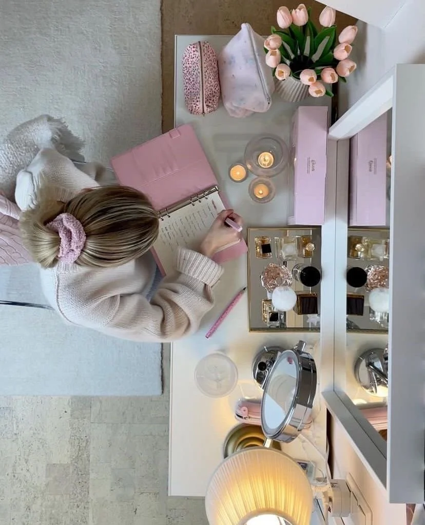 Top-down view of a student with a pink scrunchie writing in a pink planner at a white vanity desk decorated with tulips, candles, and perfumes.