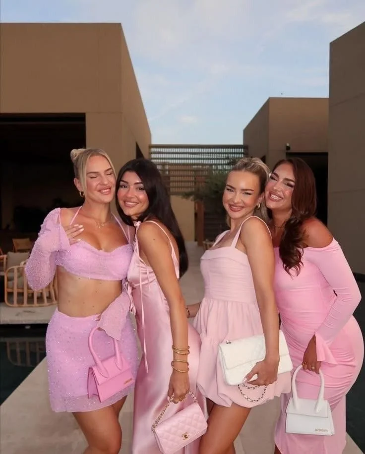 Four friends posing outdoors near a pool, wearing coordinated pink party outfits including a sequin two-piece, a satin slip dress, and mini dresses with matching handbags.