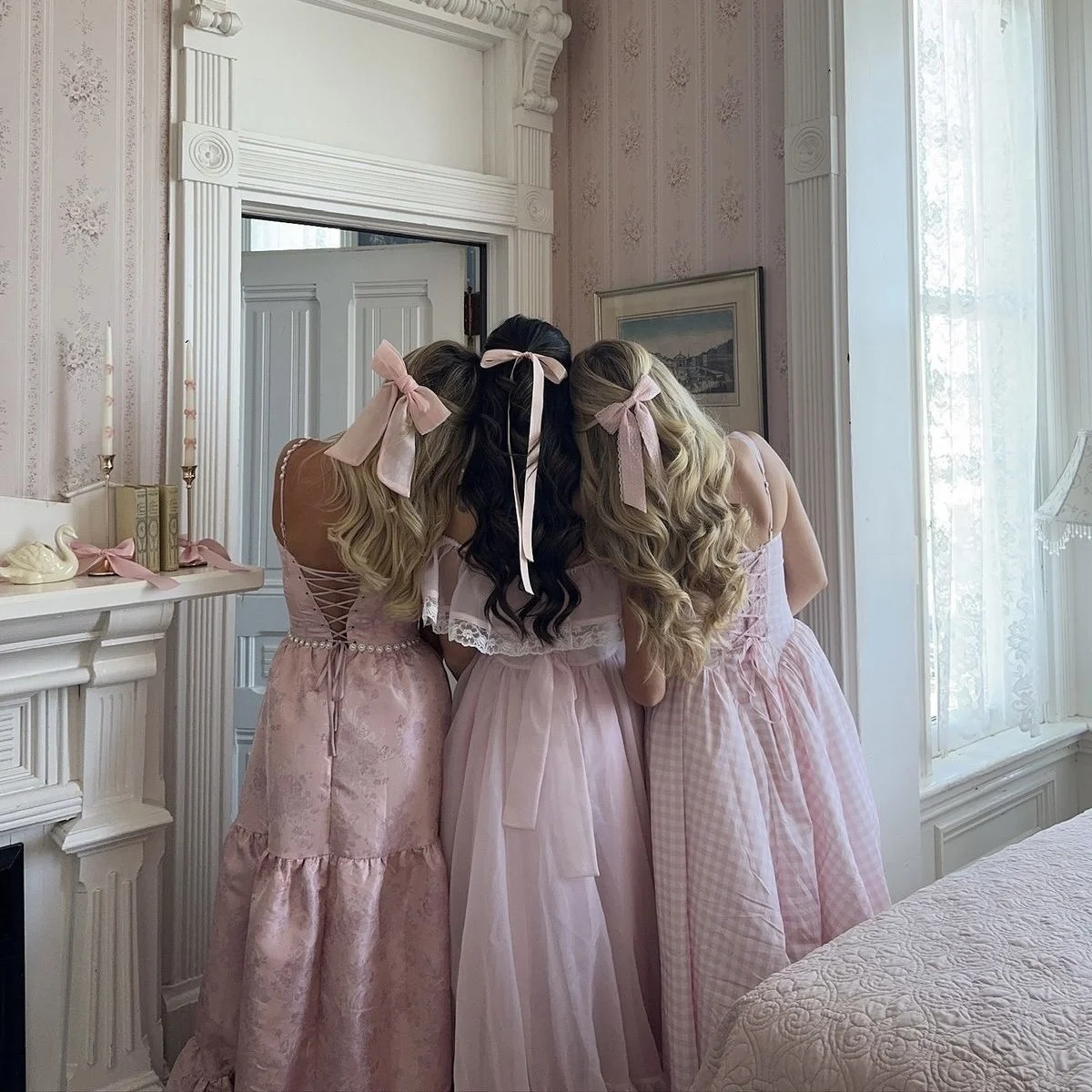 Back view of three friends hugging, wearing matching pink dresses and pink hair bows in a vintage-style room.