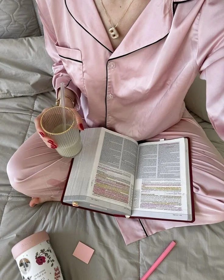 Person sitting on a bed wearing pink silk pajamas, holding a glass of iced coffee and reading a book with highlighted text.