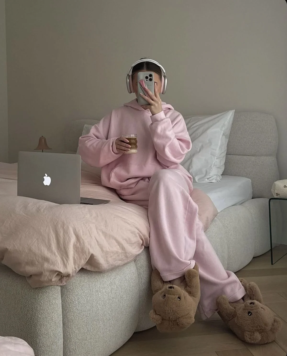 Person sitting on a bed wearing a pink hoodie, sweatpants, and teddy bear slippers, holding an iced coffee and taking a mirror selfie while working on a laptop.