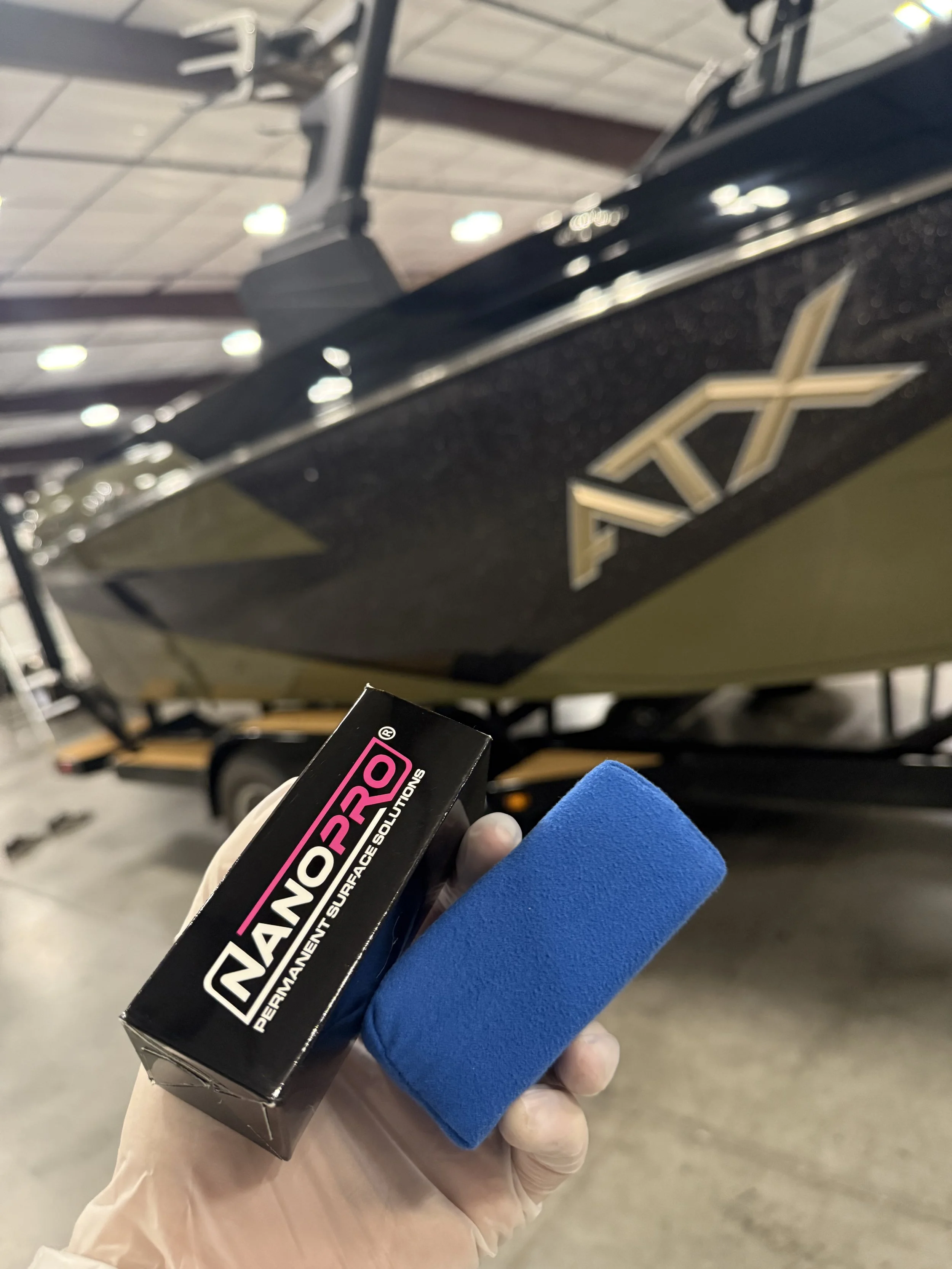 Hand holding a black box labeled 'NANOPRO' with a blue foam applicator attached, in a garage with a car in the background.