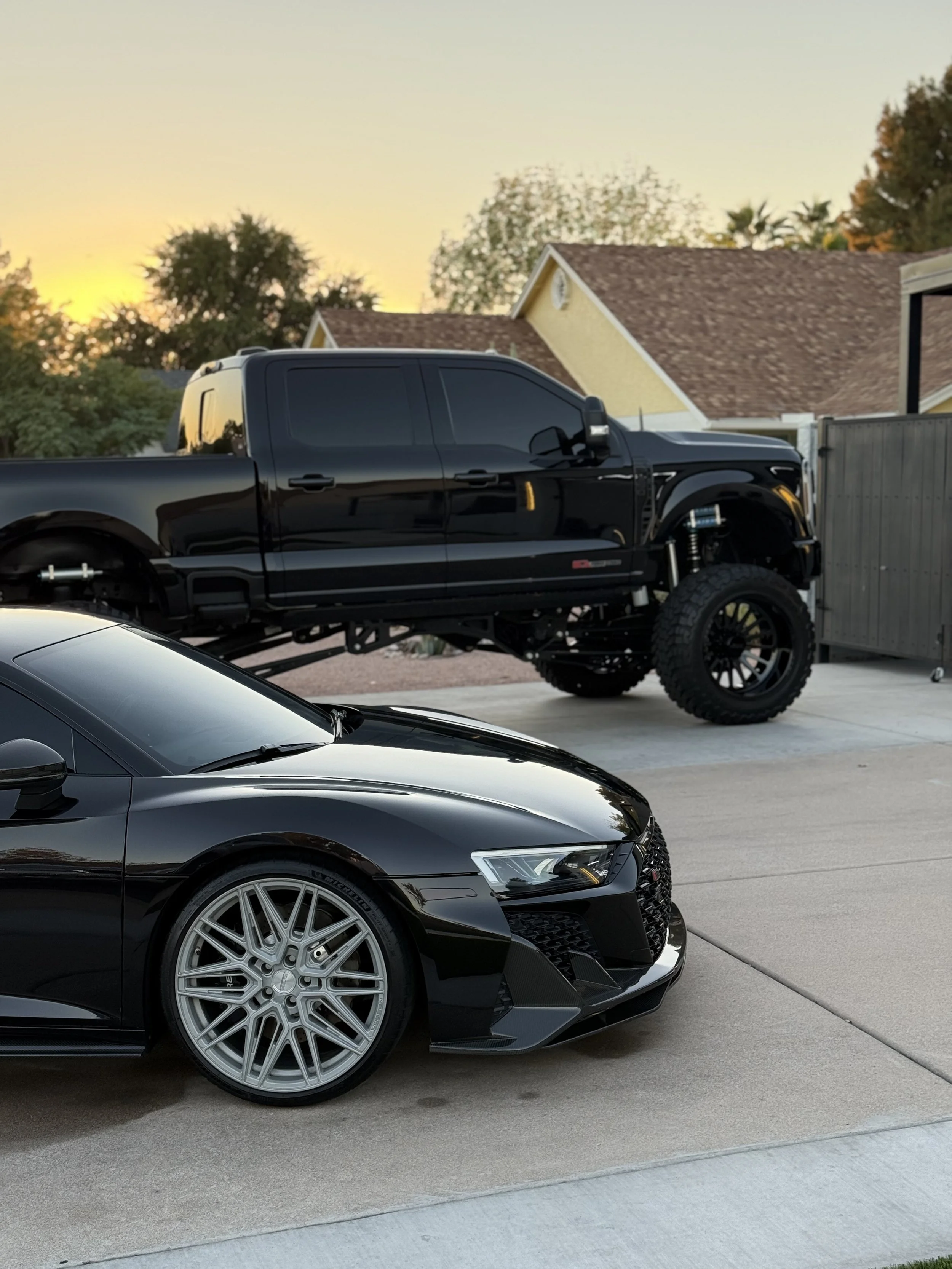 A black sports car with large silver rims parked on a driveway in front of a house with a sloped roof during sunset.