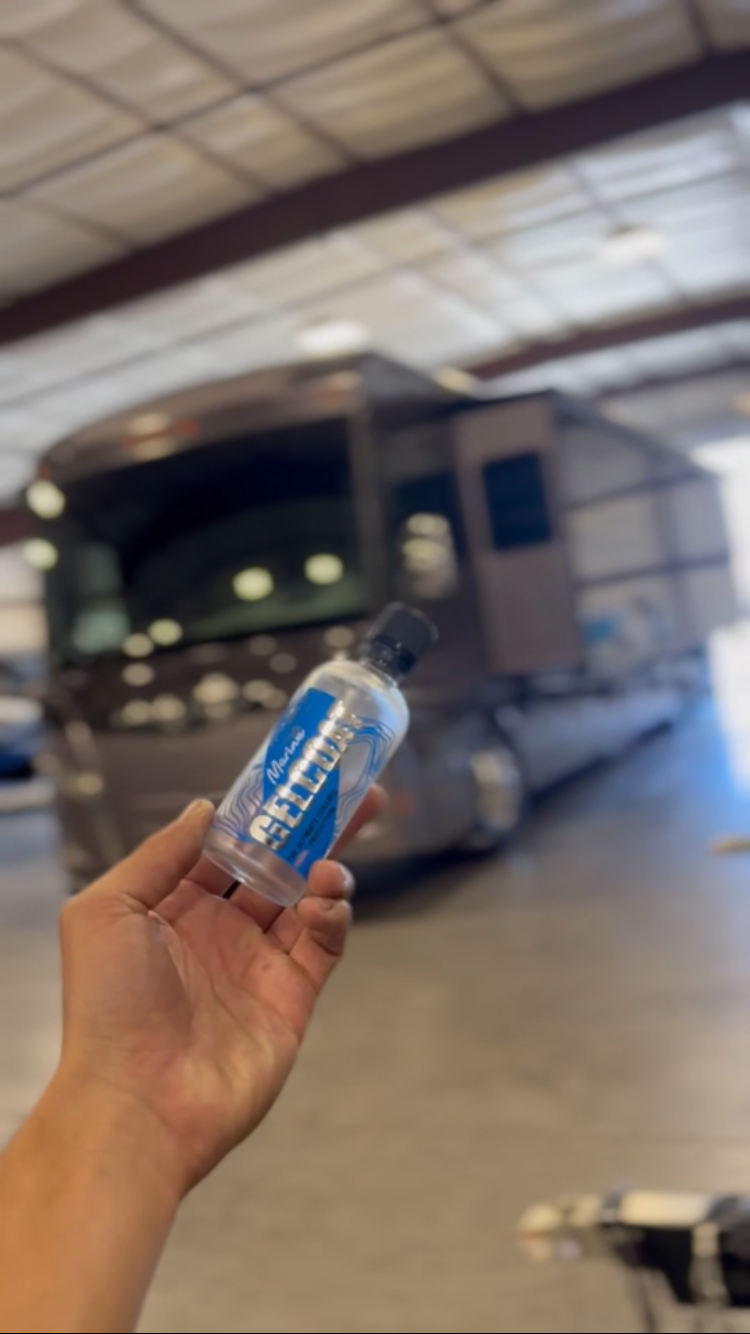 A person holding a small bottle of ceramic coating in front of a large recreational vehicle or camper in an indoor setting.