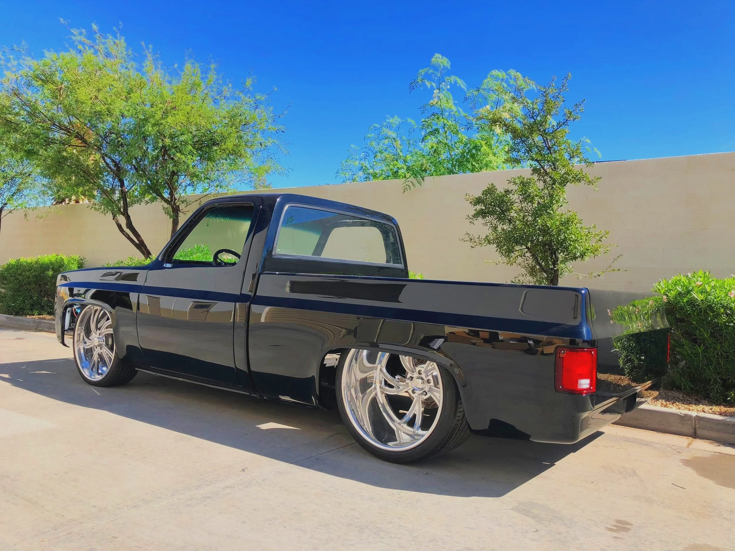 A black pickup truck with large, shiny aftermarket wheels parked on a concrete surface in front of a beige wall and green shrubs. Behind the wall, there are multiple green trees under a bright blue sky.
