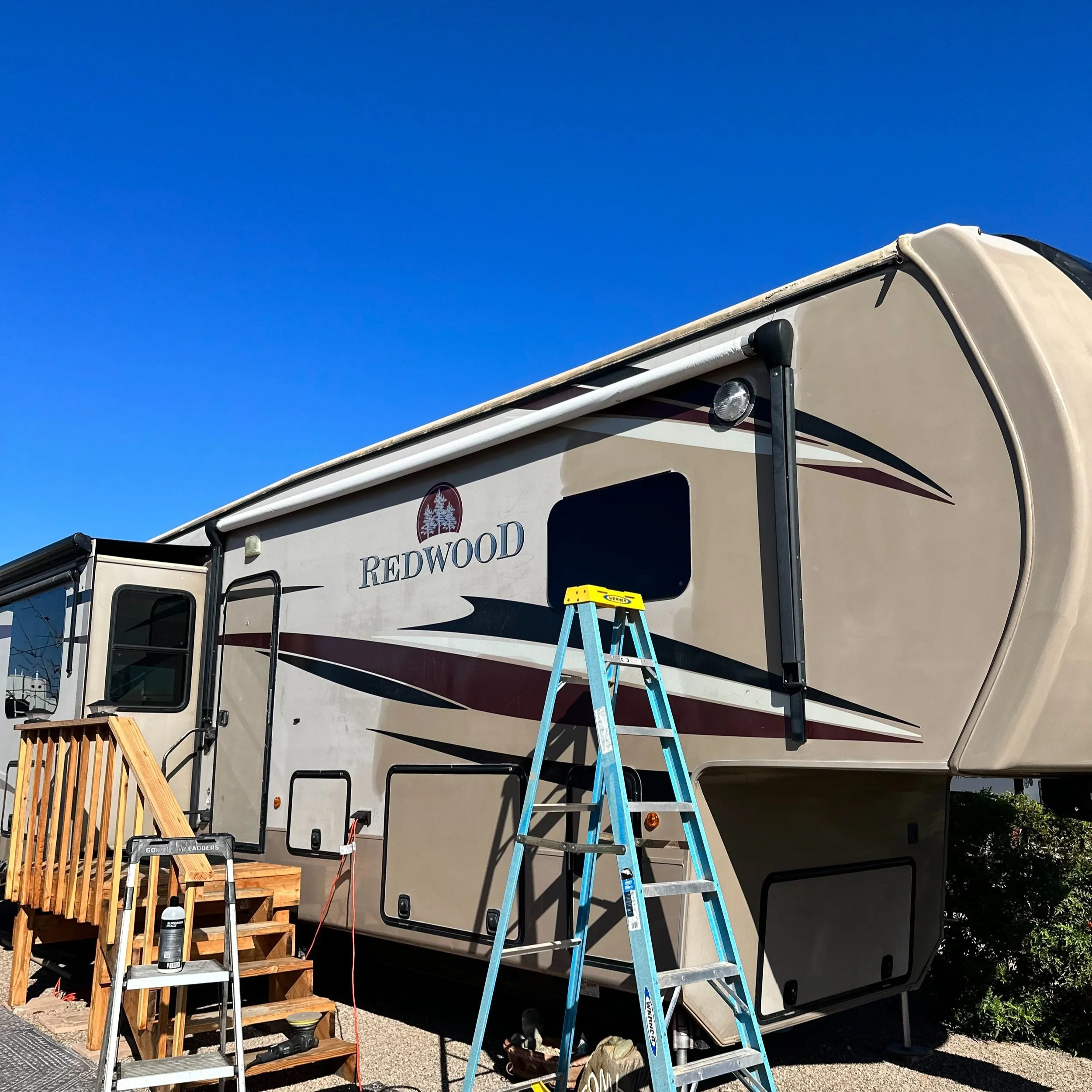 Working on exterior of Winnebago RV with ladders, wooden furniture, tools, and cleaning supplies nearby, under a clear blue sky.