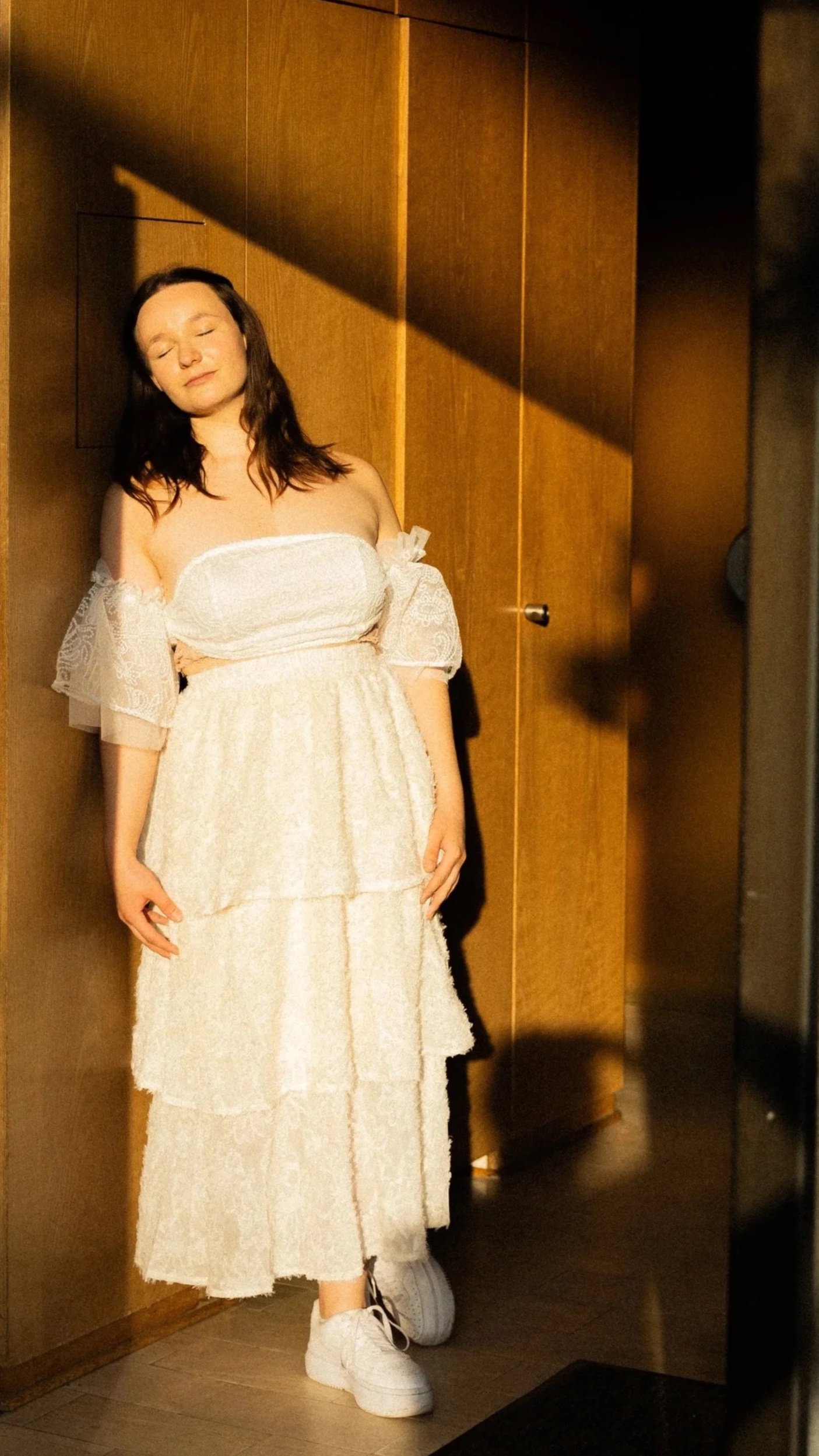 Junge Frau in einem weißen, mehrlagigen Kleid mit Spitzen-Details steht entspannt an einer Holzwand, die ihr im Sonnenlicht warm leuchtet.