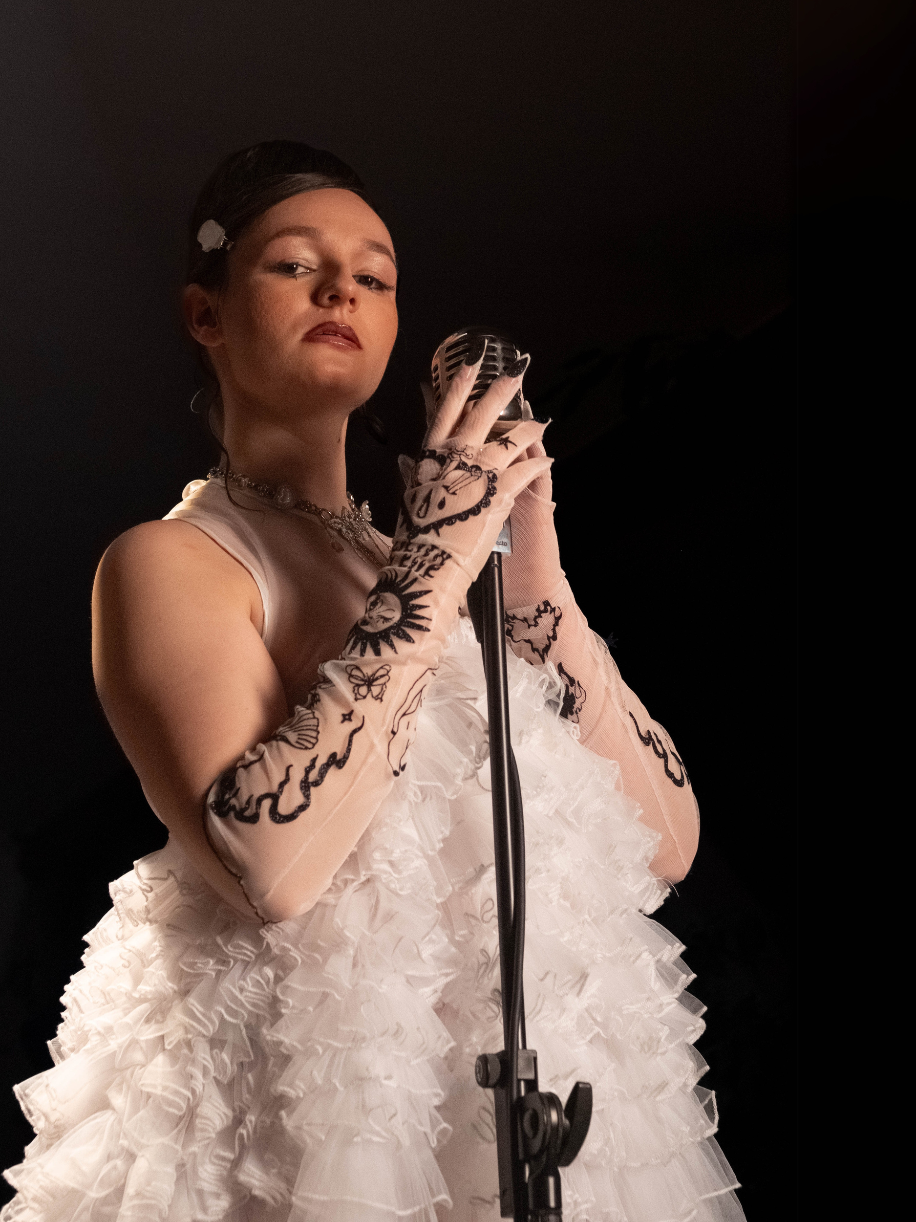 Junge Frau in einem weiß gewellten Kleid und Spitzenhandschuhen steht vor einem Mikrofon, schaut selbstbewusst in die Kamera.