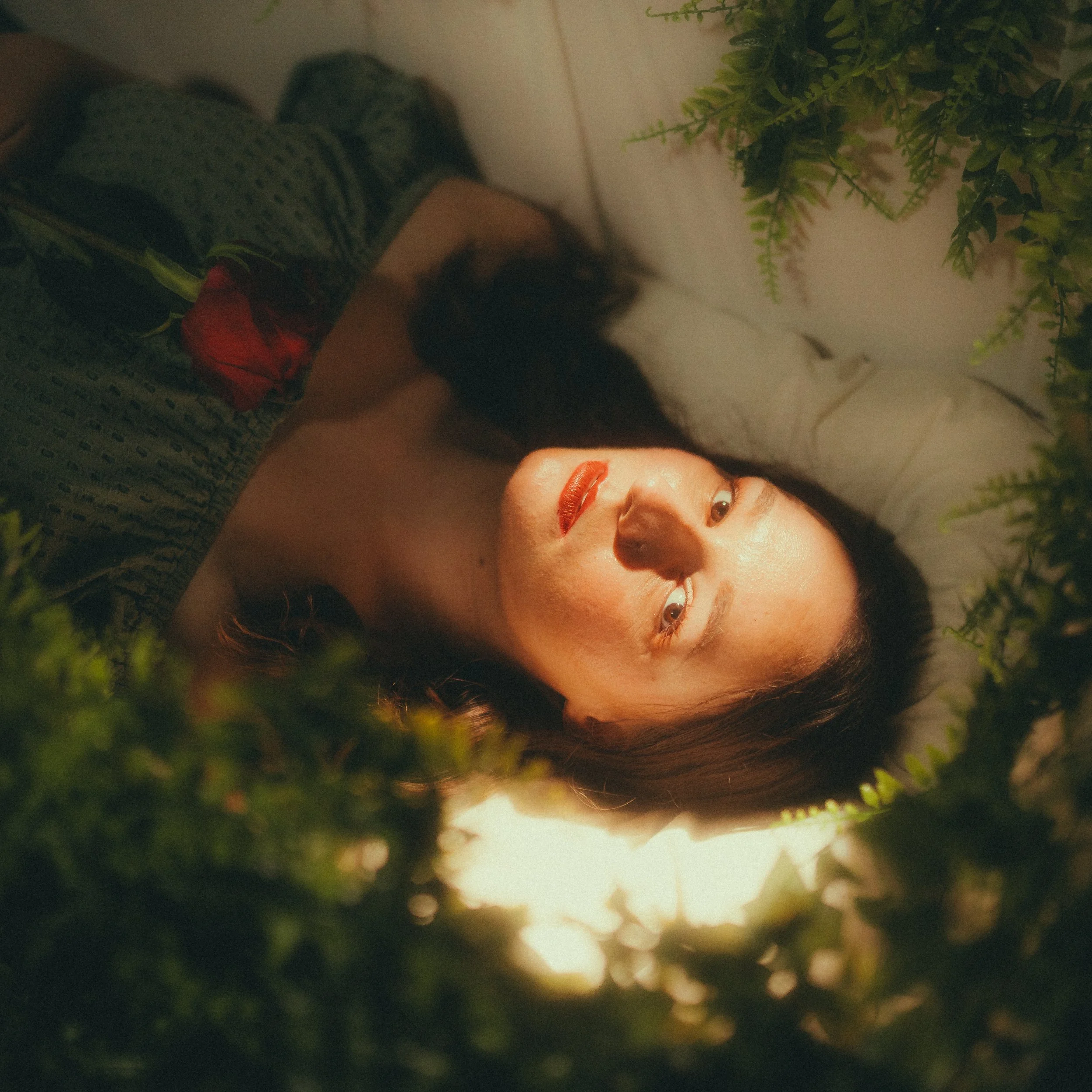 Frau mit dunklen Haaren liegt auf dem Rücken, umgeben von grünen Blättern, und schaut zur Seite. Sie hält eine einzelne rosafarbene Rose in ihrer Hand.