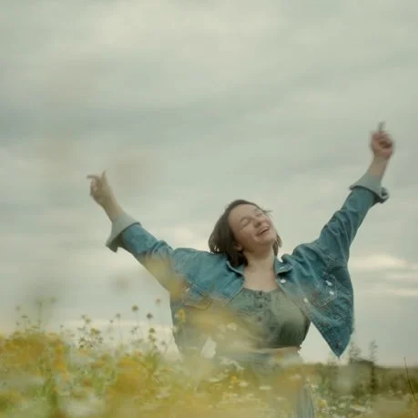 Eine junge Frau steht in einem Feld mit ihren Armen hoch in der Luft und lächelt bei sonnigem Himmel.