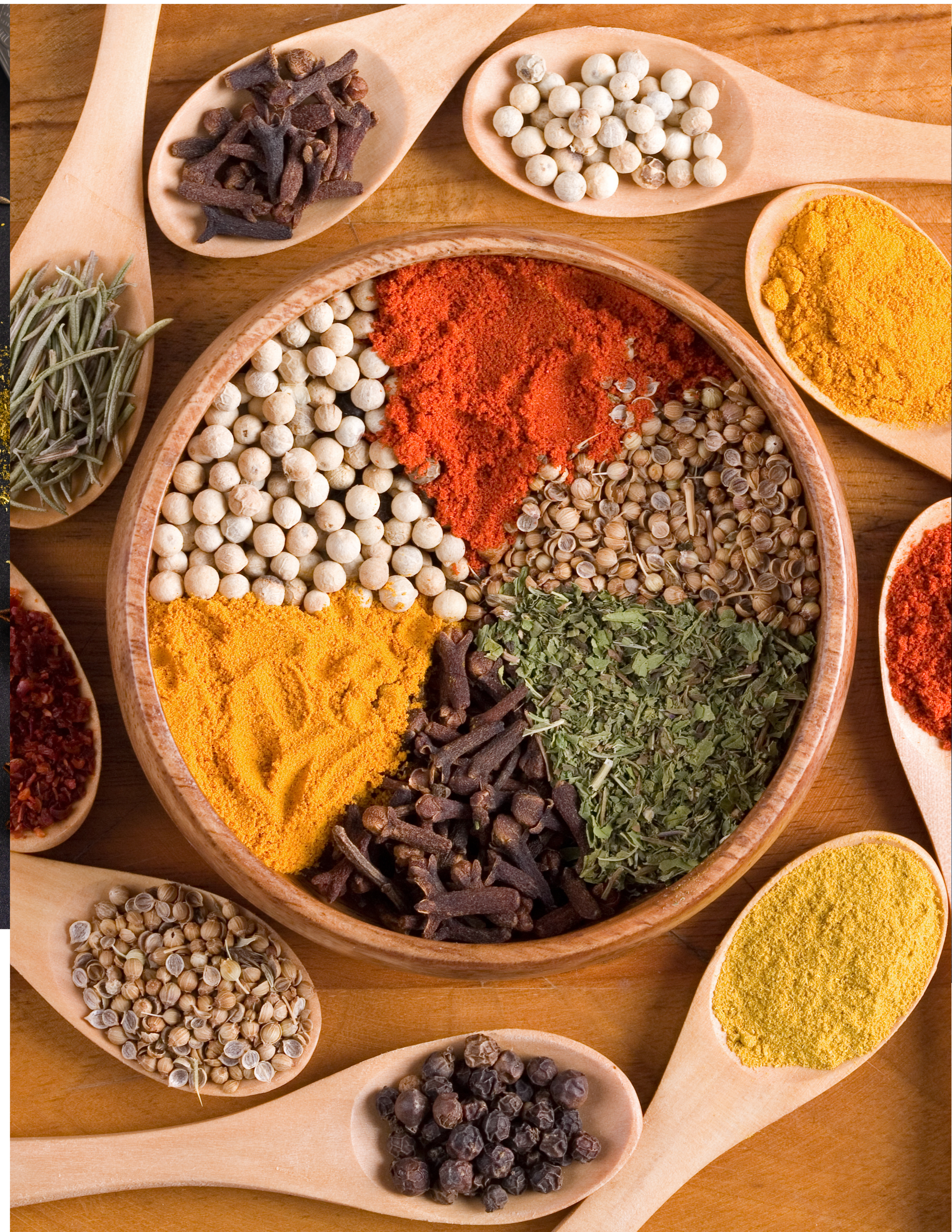 Various spices in wooden spoons and a round wooden bowl, including white peppercorns, turmeric, chili powder, dried herbs, and black pepper, on a wooden surface.