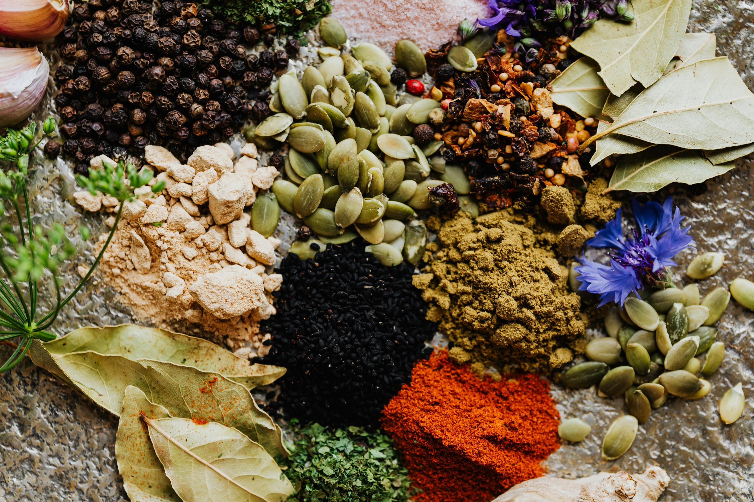Various spices and herbs arranged on a surface, including black peppercorns, pumpkin seeds, turmeric powder, dried bay leaves, dried chili powder, dried herbs, dried onion flakes, and dried flower.