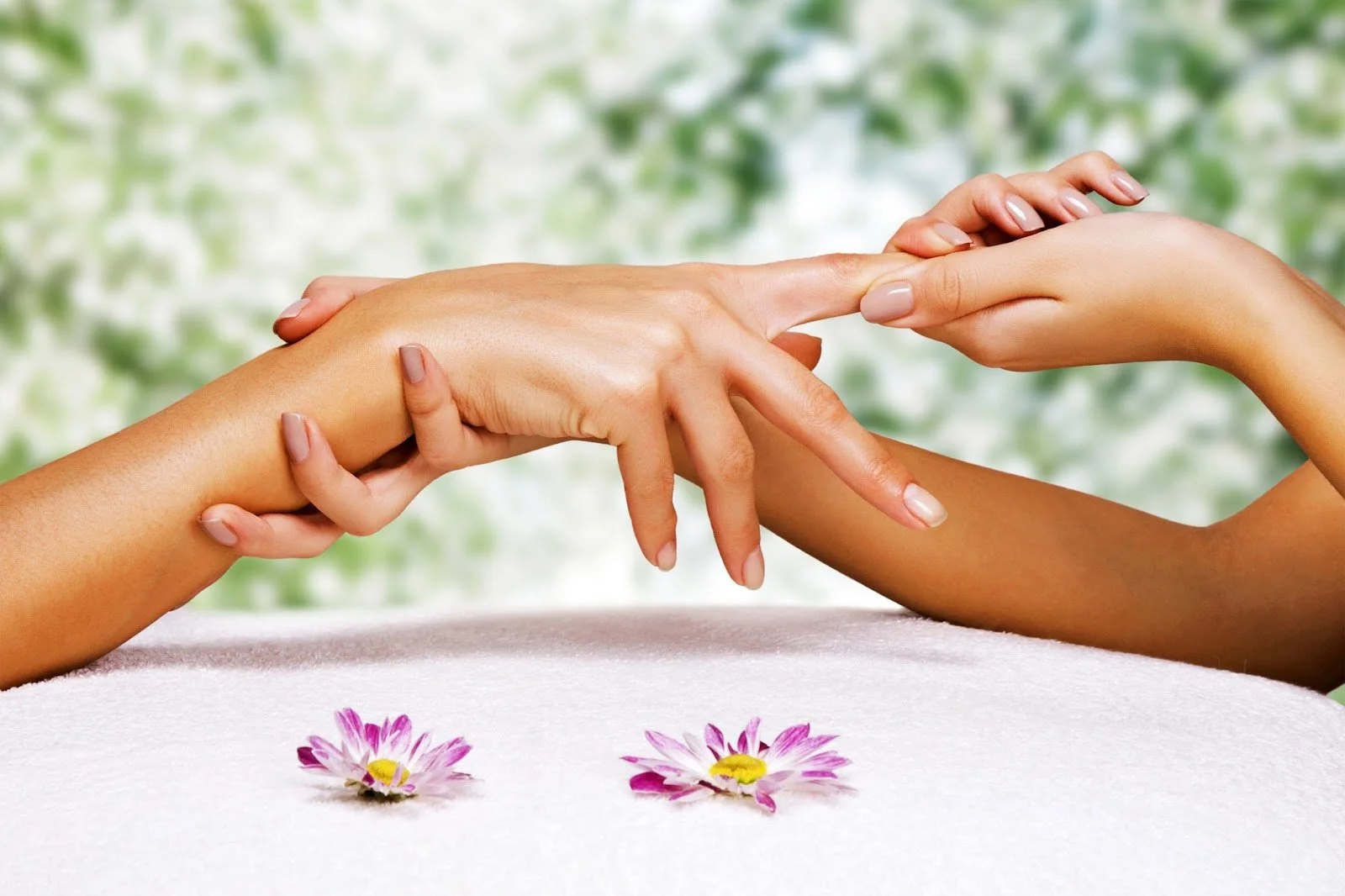 A hand receiving a massage while another hand applies the massage, with pink and white flowers on a white surface and a green blurred background.