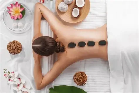 A woman receiving a hot stone massage, with four black stones on her back, surrounded by spa items like flowers, candles, and decor.