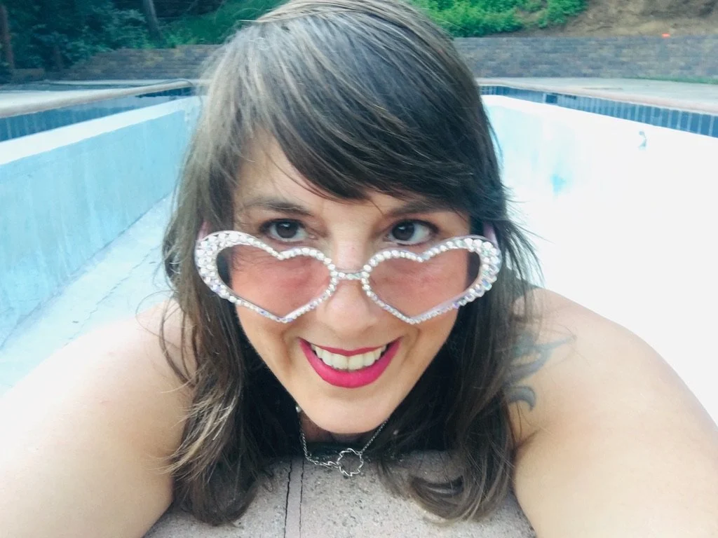 Close-up of a woman with brown hair, wearing a floral blouse and pearl necklace, smiling with rose colored heart sunglasses