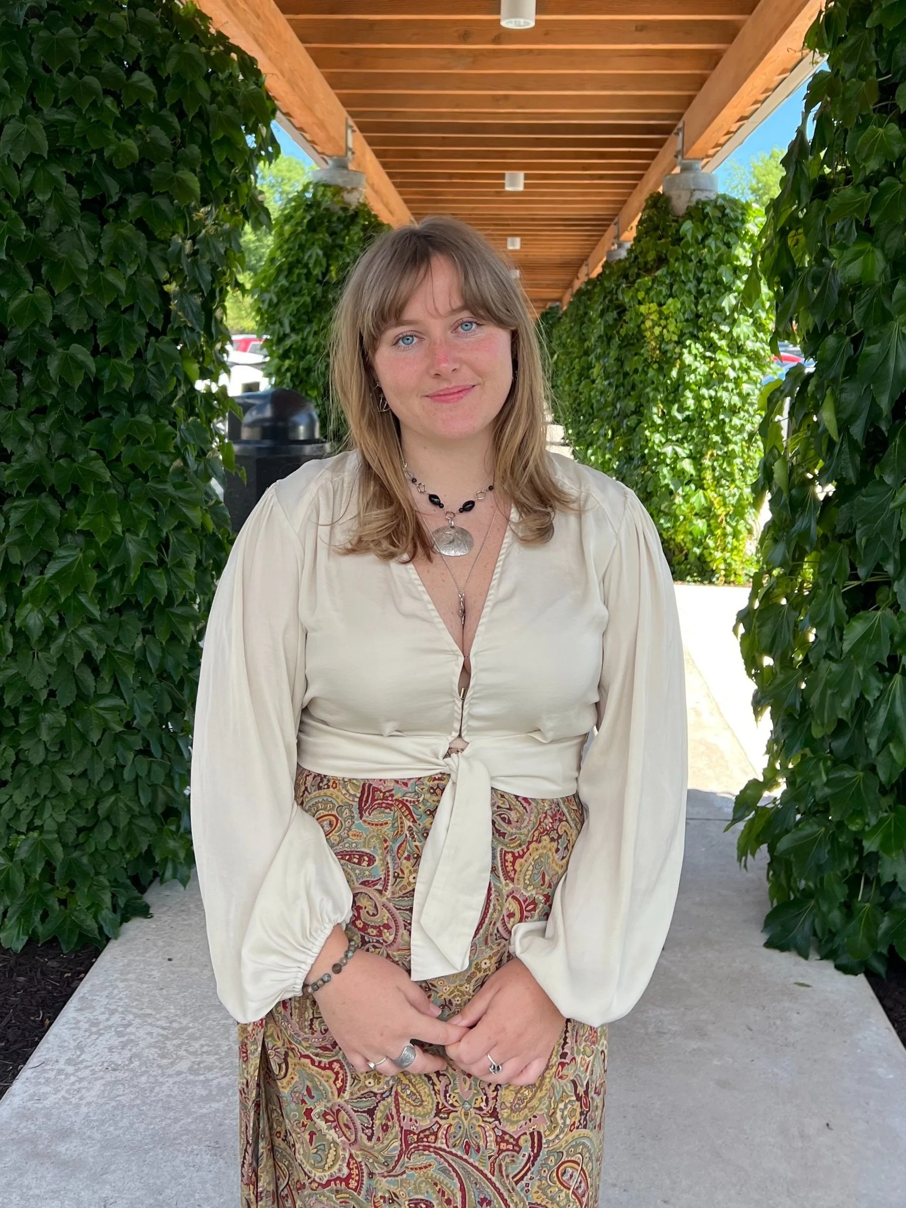 Woman in sunglasses holding a bouquet of flowers on a sandy beach with green grass and a water tower in the background during sunset.