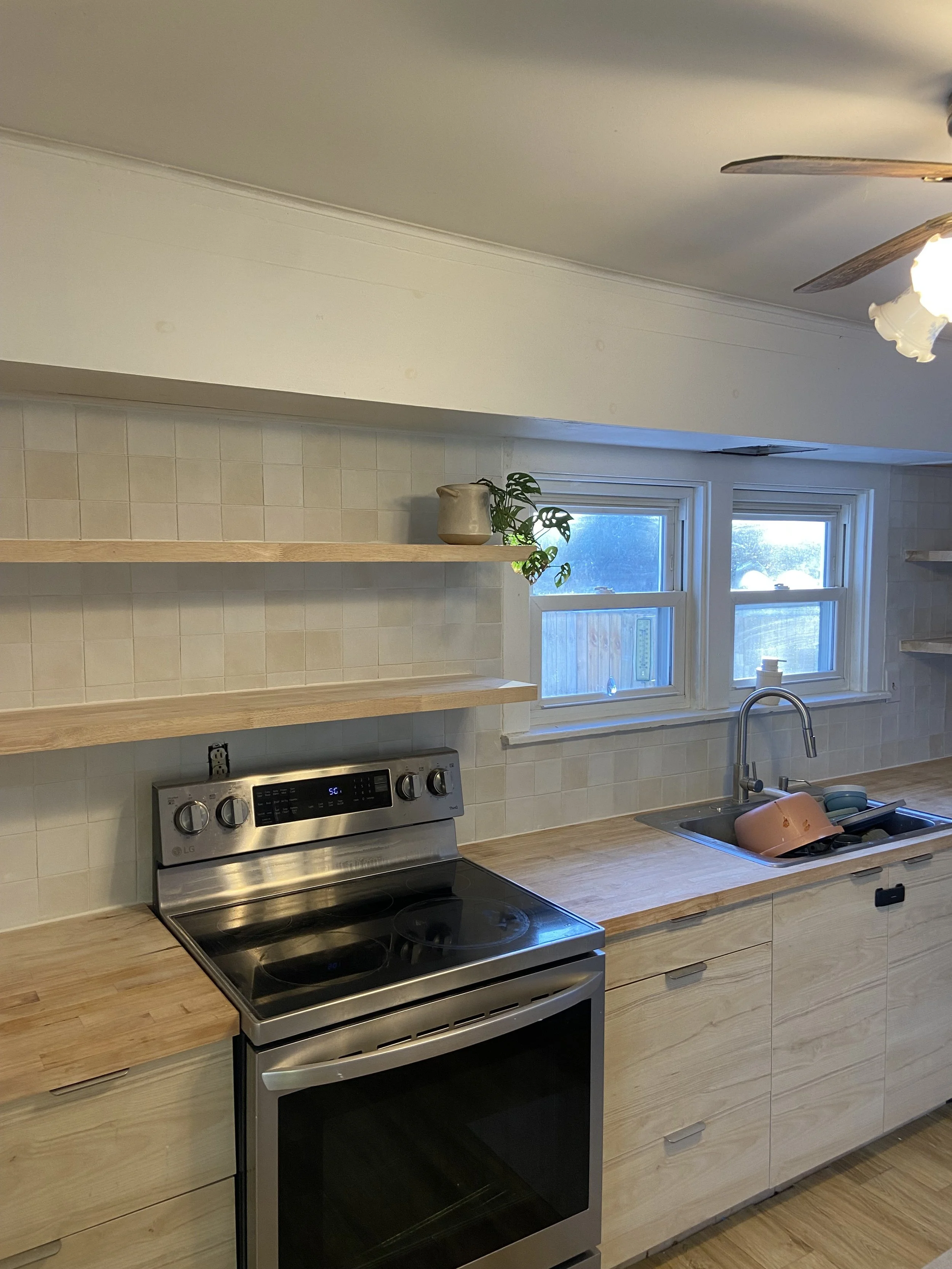 Modern kitchen with stainless steel stove, wooden cabinets, open shelves, a sink with dishes, and a ceiling fan.