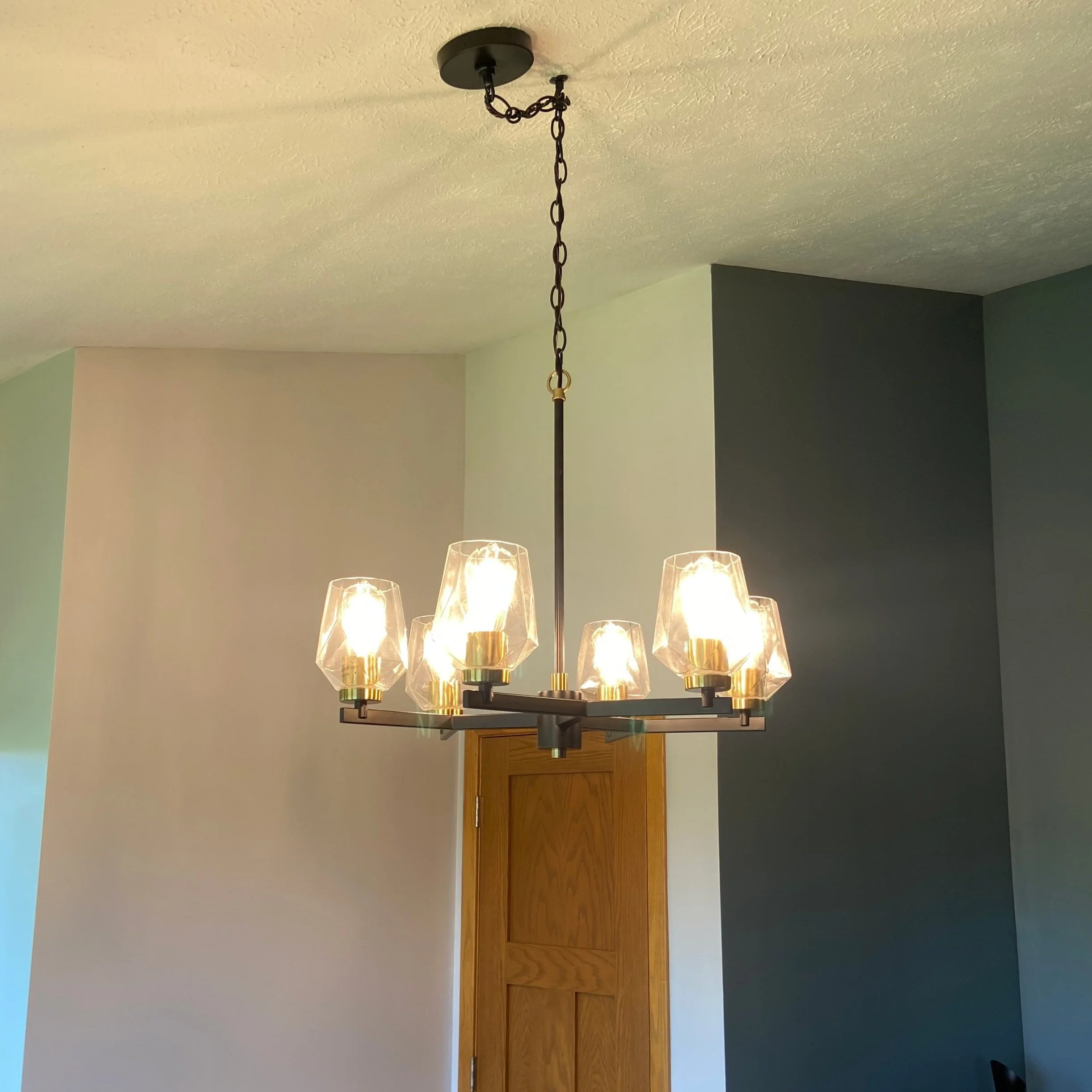 A modern chandelier with six glass shades hanging from a black metal frame, illuminated and hanging from a chain attached to the ceiling.