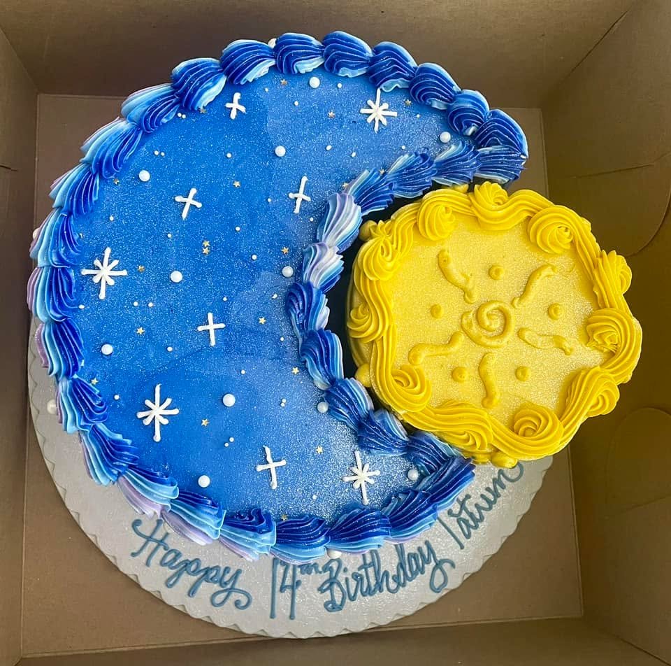 A large birthday cake with a starry night sky theme on one side and a yellow clock face on the other, decorated with blue and purple icing, surrounded by a white base with a birthday message.