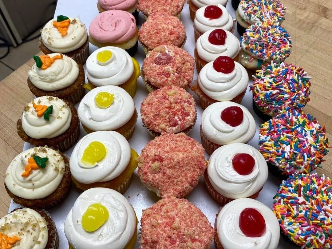 Assorted cupcakes decorated with various colorful toppings, including sprinkles, cherries, lemon, and carrot-shaped decorations.