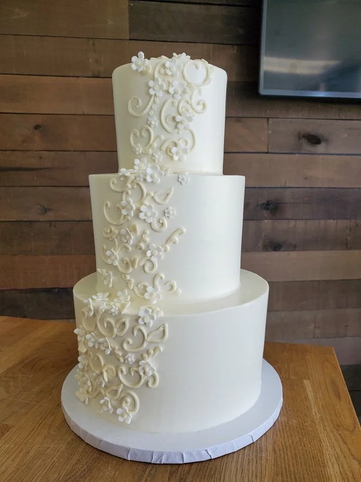 A three-tiered white wedding cake decorated with intricate floral and scroll patterns, placed on a wooden table.