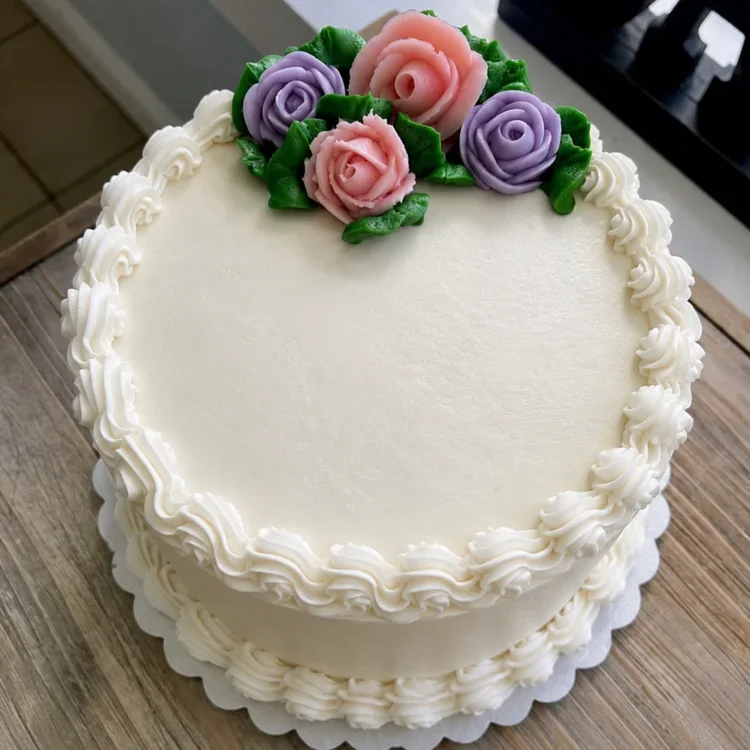 Round white cake decorated with piped white frosting border and pink and purple buttercream roses on top, placed on a wooden surface.
